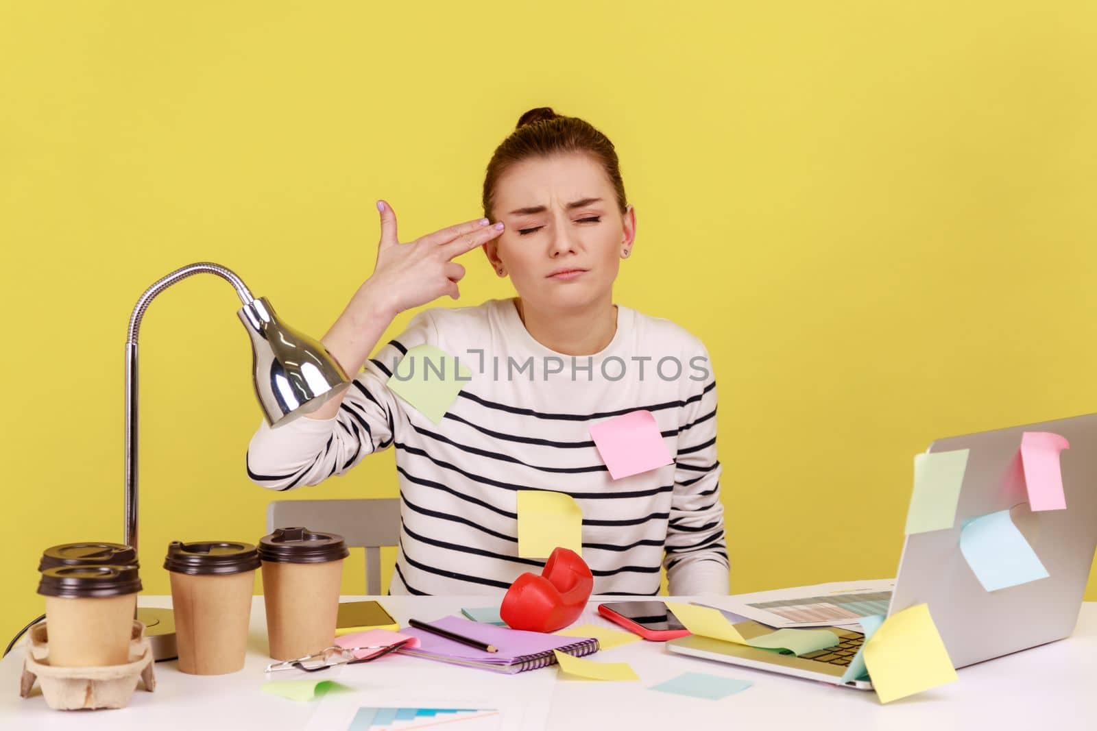 Depressed woman covered with sticky notes working on laptop, pointing finger gun gesture to head, committing suicide from stressful job. Indoor studio studio shot isolated on yellow background.