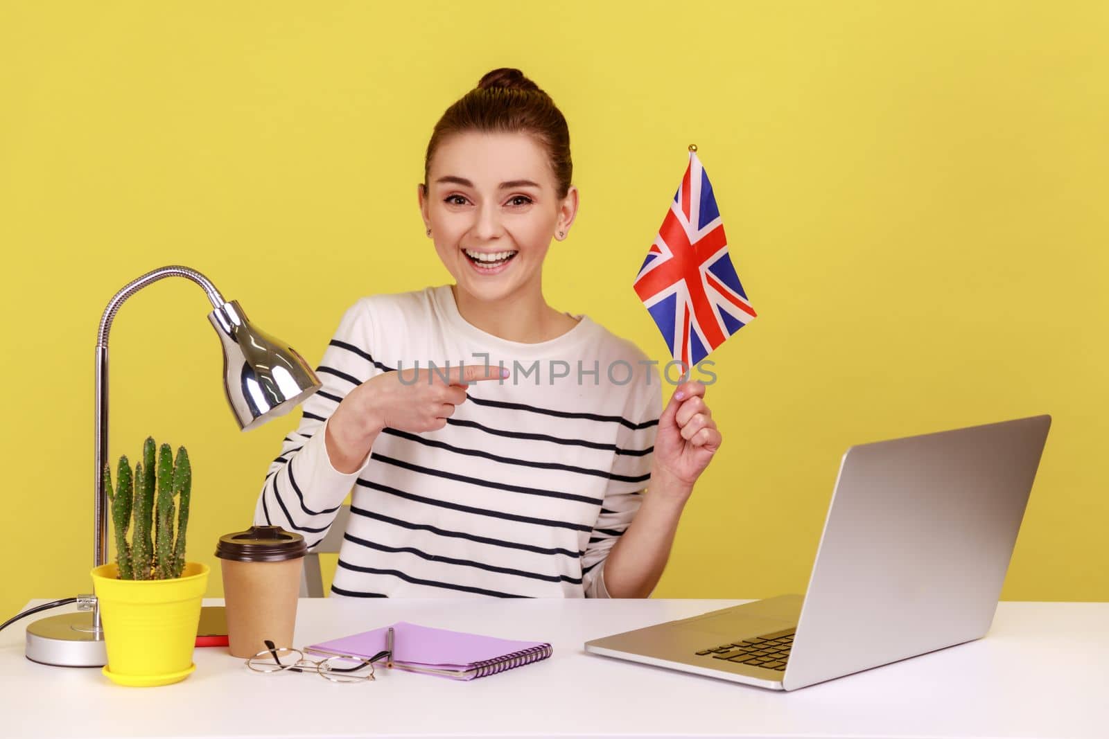 Smiling positive woman sitting at workplace with, pointing at flag of United Kingdom of Great Britain, expressing happiness, relocation abroad. Indoor studio studio shot isolated on yellow background.