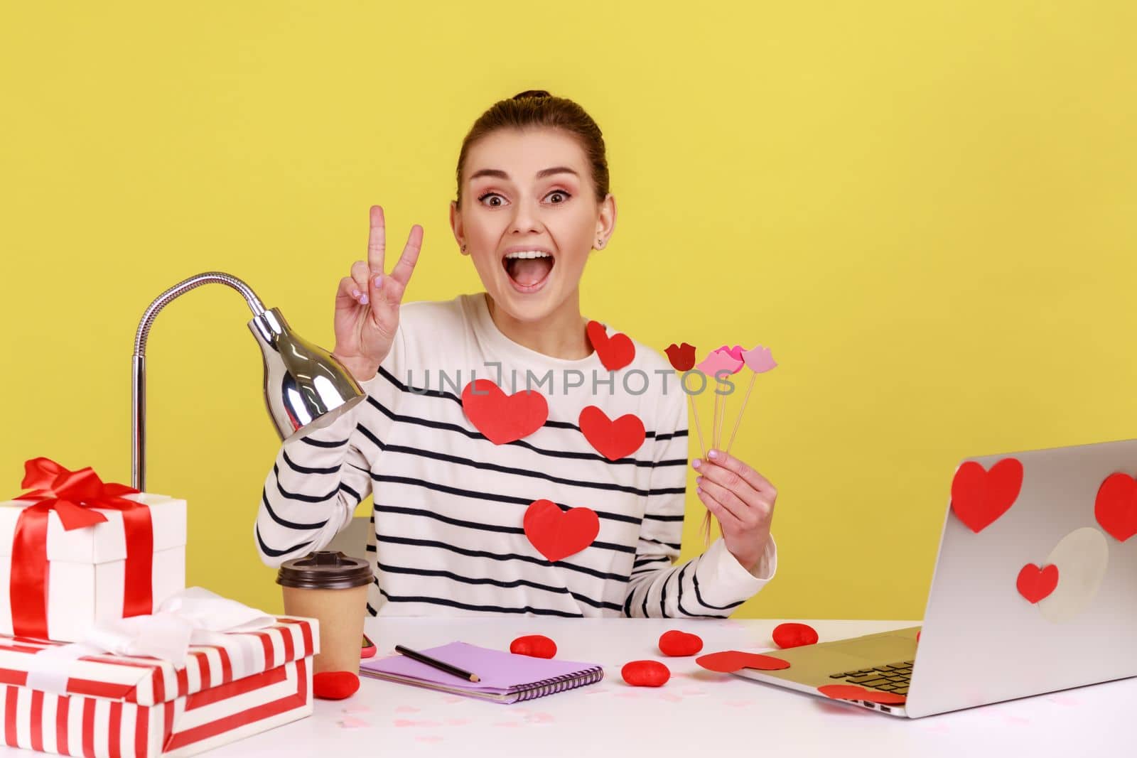 Funny excited festive woman office manager sitting on workplace with pout lips, holding party props, showing v sign, looking at camera. Indoor studio studio shot isolated on yellow background.