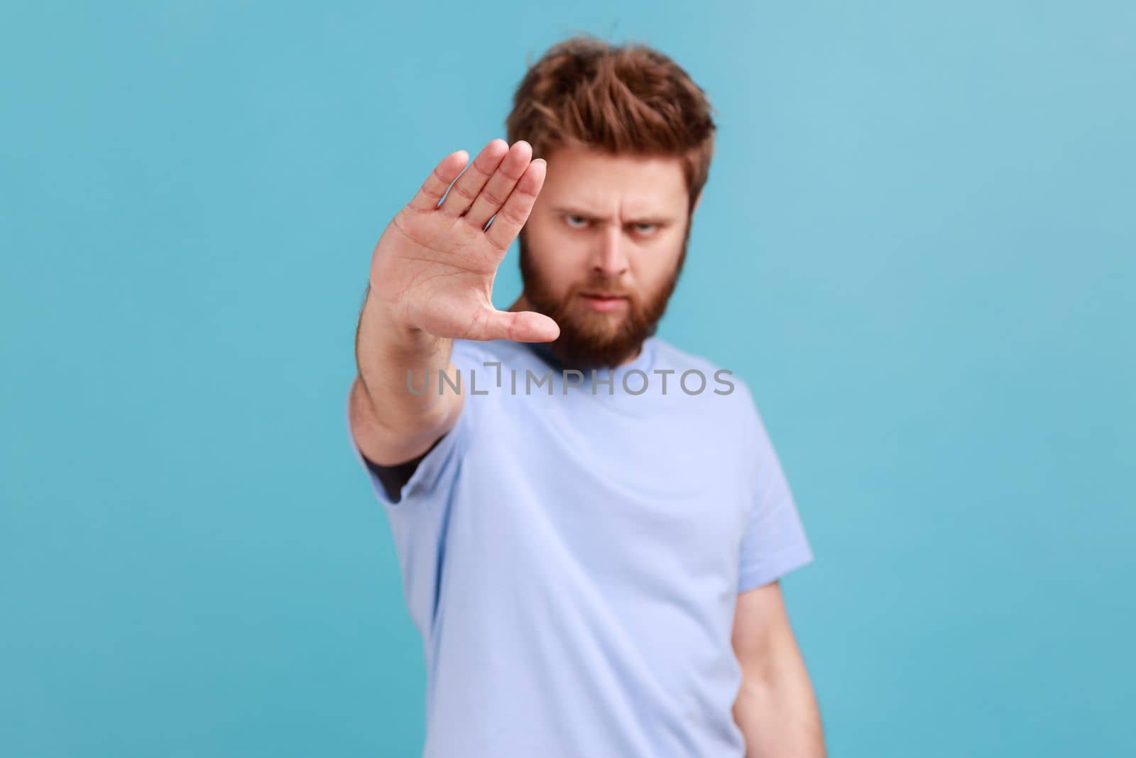 Portrait of strict bearded man showing stop sign at camera, standing with frowning face and bossy expression, meaning caution to avoid danger or mistake. Indoor studio shot isolated on blue background