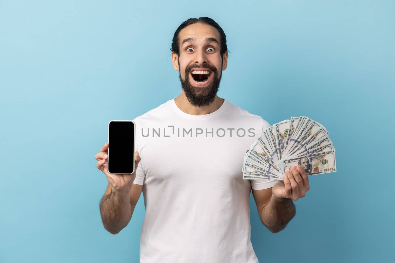 Portrait of man with beard wearing white T-shirt holding fan of dollar banknotes and smart phone with black empty display, making money online. Indoor studio shot isolated on blue background.