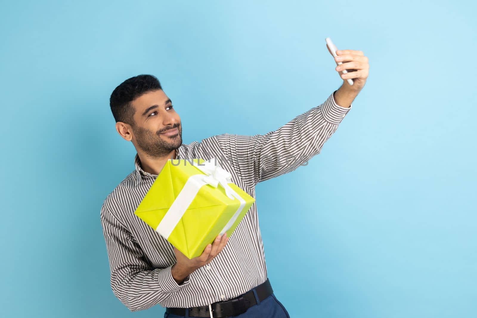 Portrait of positive bearded businessman making selfie or recording video, famous blogger preparing gifts for subscribers, wearing striped shirt. Indoor studio shot isolated on blue background.