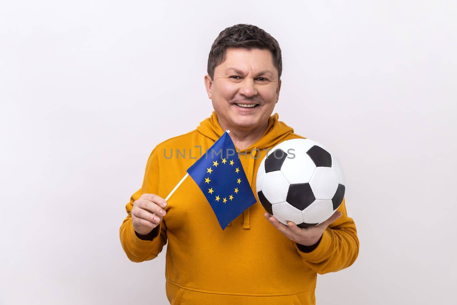 Satisfied middle aged man holds in hands flag of europe union and football ball, supporting favourite team on championship, wearing urban style hoodie. Indoor studio shot isolated on white background.