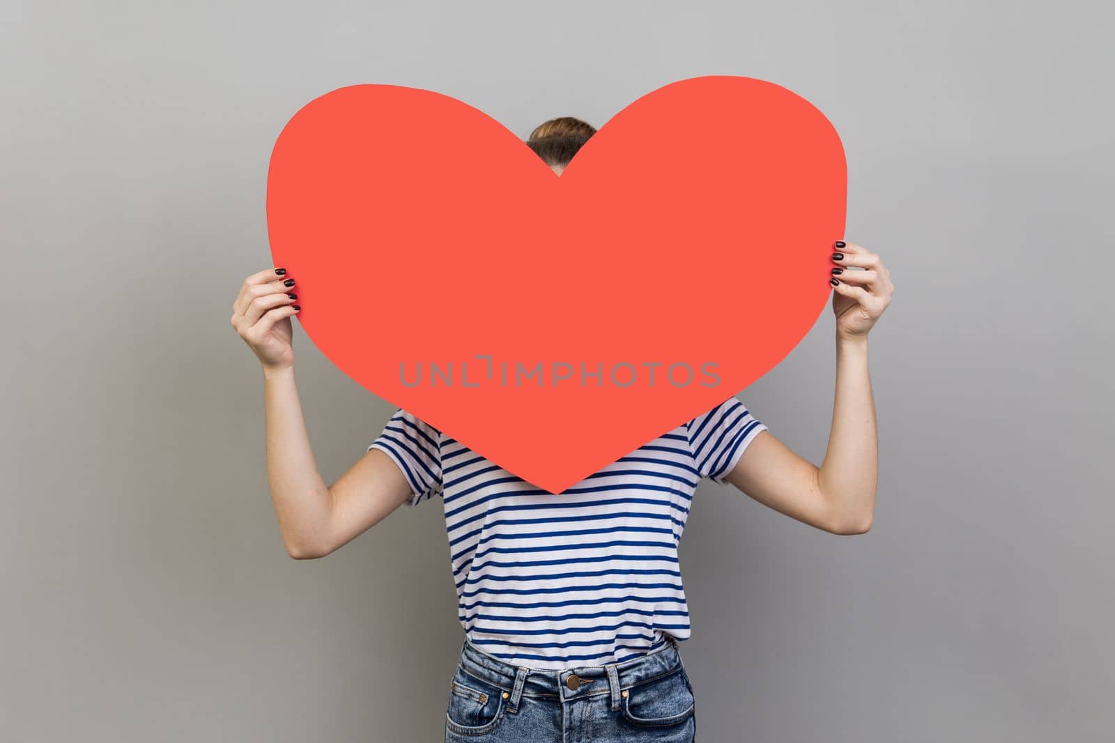 Portrait of unknown unrecognizable woman wearing striped T-shirt hiding her face behind big red heart, hesitates to confess his feelings. Indoor studio shot isolated on gray background.