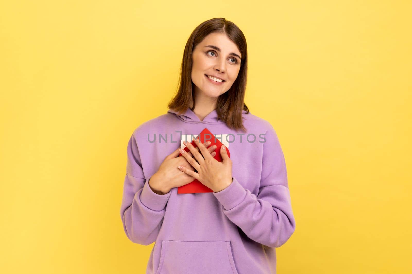 Portrait of beautiful woman standing, embracing envelope and looking away with pleasant smile and enjoying, wearing purple hoodie. Indoor studio shot isolated on yellow background.