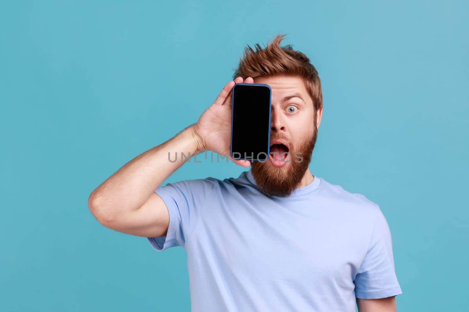 Man in T-shirt covering half face with cellphone and looking at camera with open mouth, shocked face by Khosro1