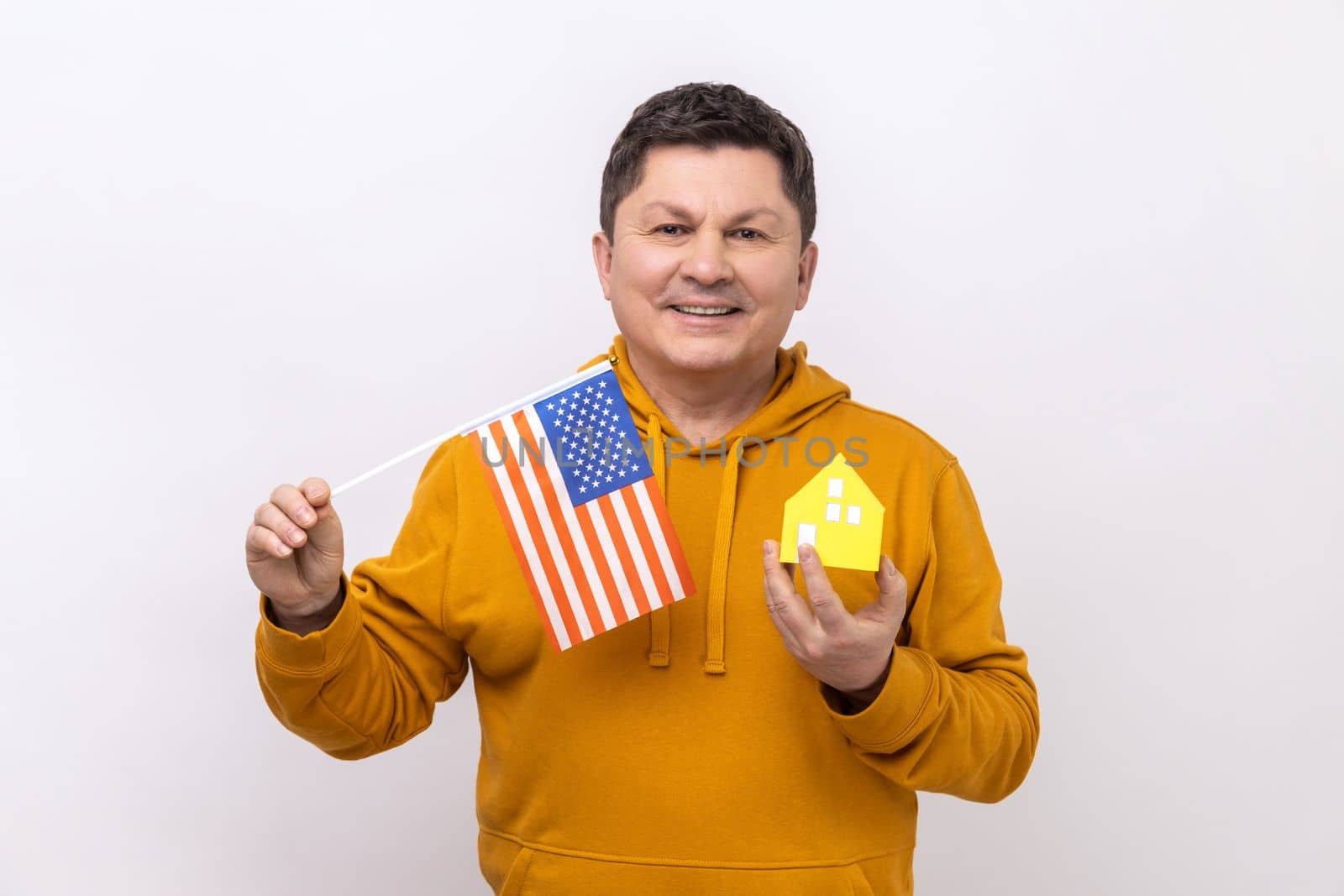 Portrait of middle aged man holding american flag and paper house, dreaming to buy accommodation in USA, wearing urban style hoodie. Indoor studio shot isolated on white background.