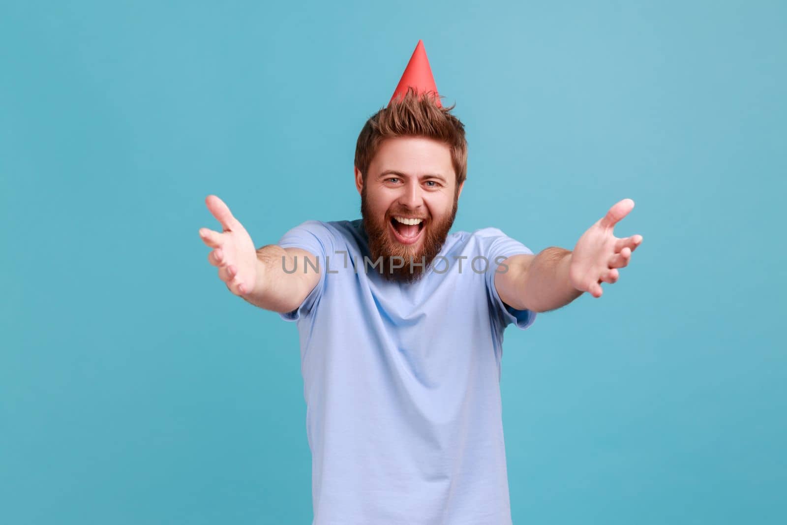 Portrait of extremely happy bearded man wearing party cone giving free hugs with outstretched hands, welcoming inviting to embrace. Indoor studio shot isolated on blue background.