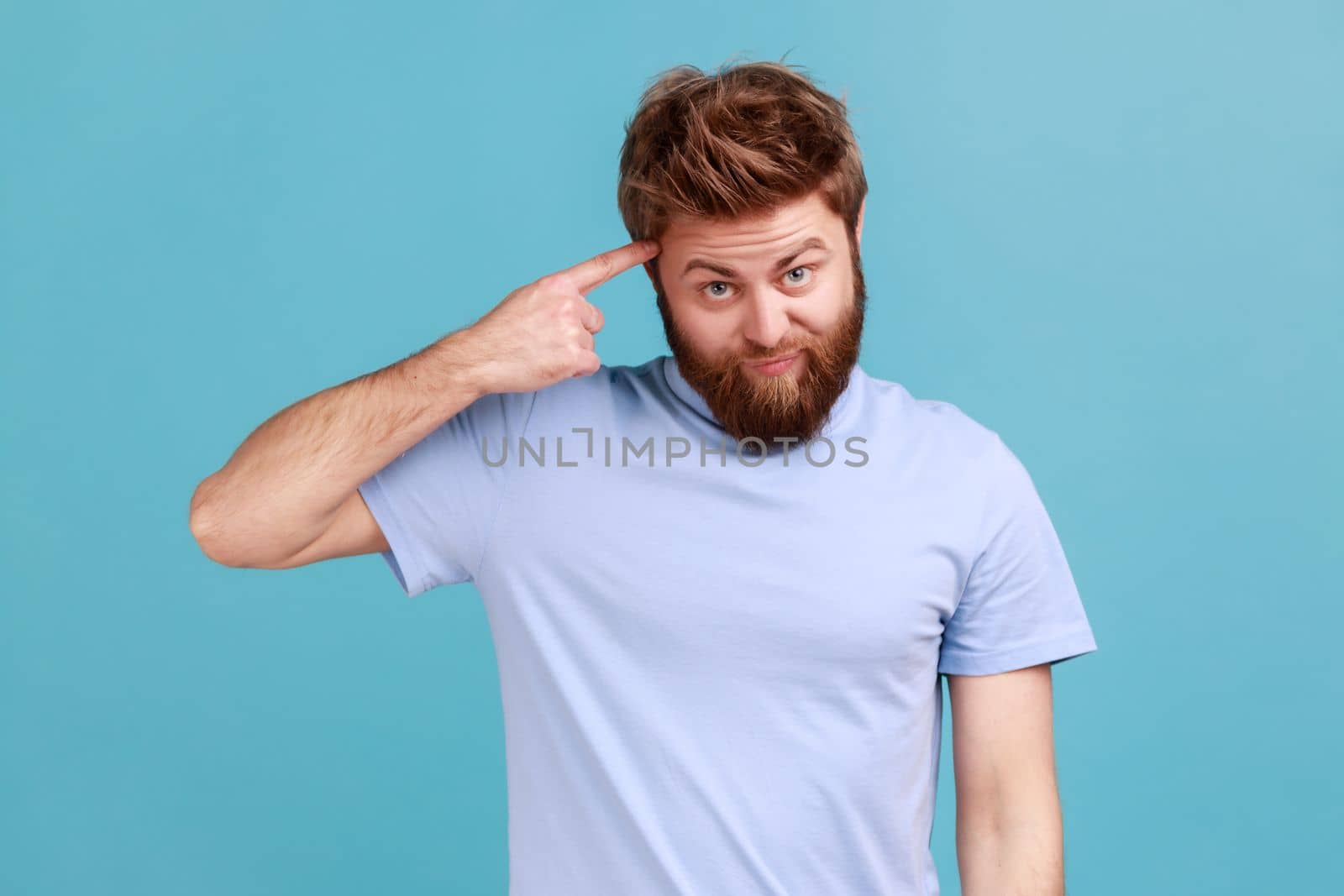 Portrait of young bearded man showing stupid gesture with finger near head, out of mind, accusing crazy dumb plan, reckless expression. Indoor studio shot isolated on blue background.
