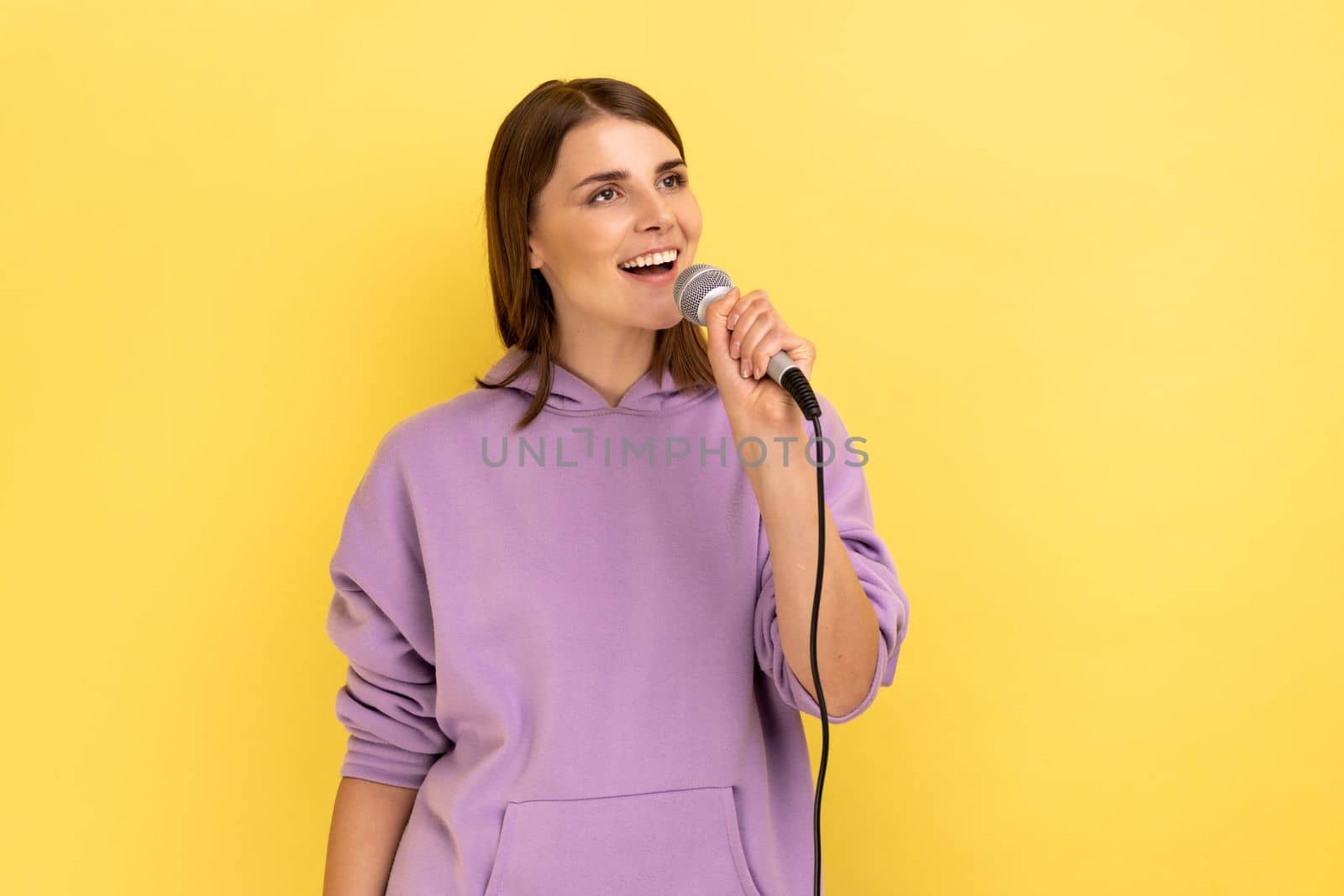Portrait of positive woman loudly singing song, holding microphone in hand, having fun resting in karaoke, singer, wearing purple hoodie. Indoor studio shot isolated on yellow background.