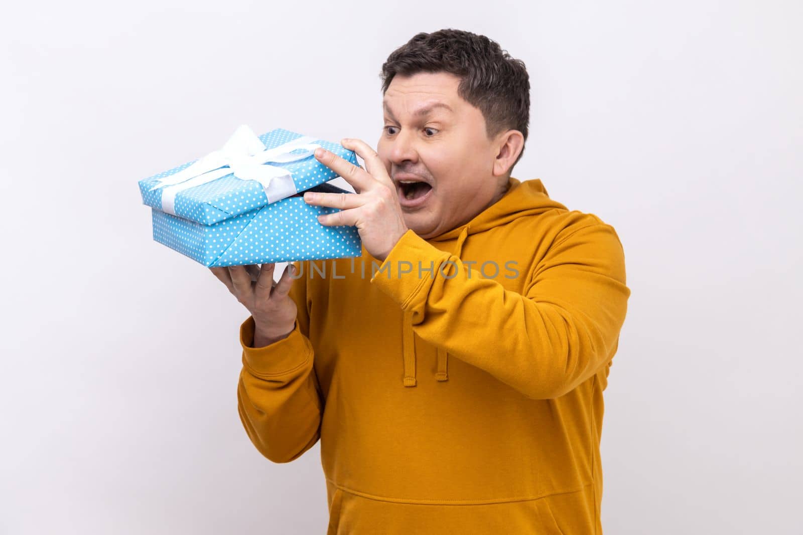 Portrait of curious middle aged man with present in hands looking inside gift box with surprised expression, unpacking gift, wearing urban style hoodie. Indoor studio shot isolated on white background