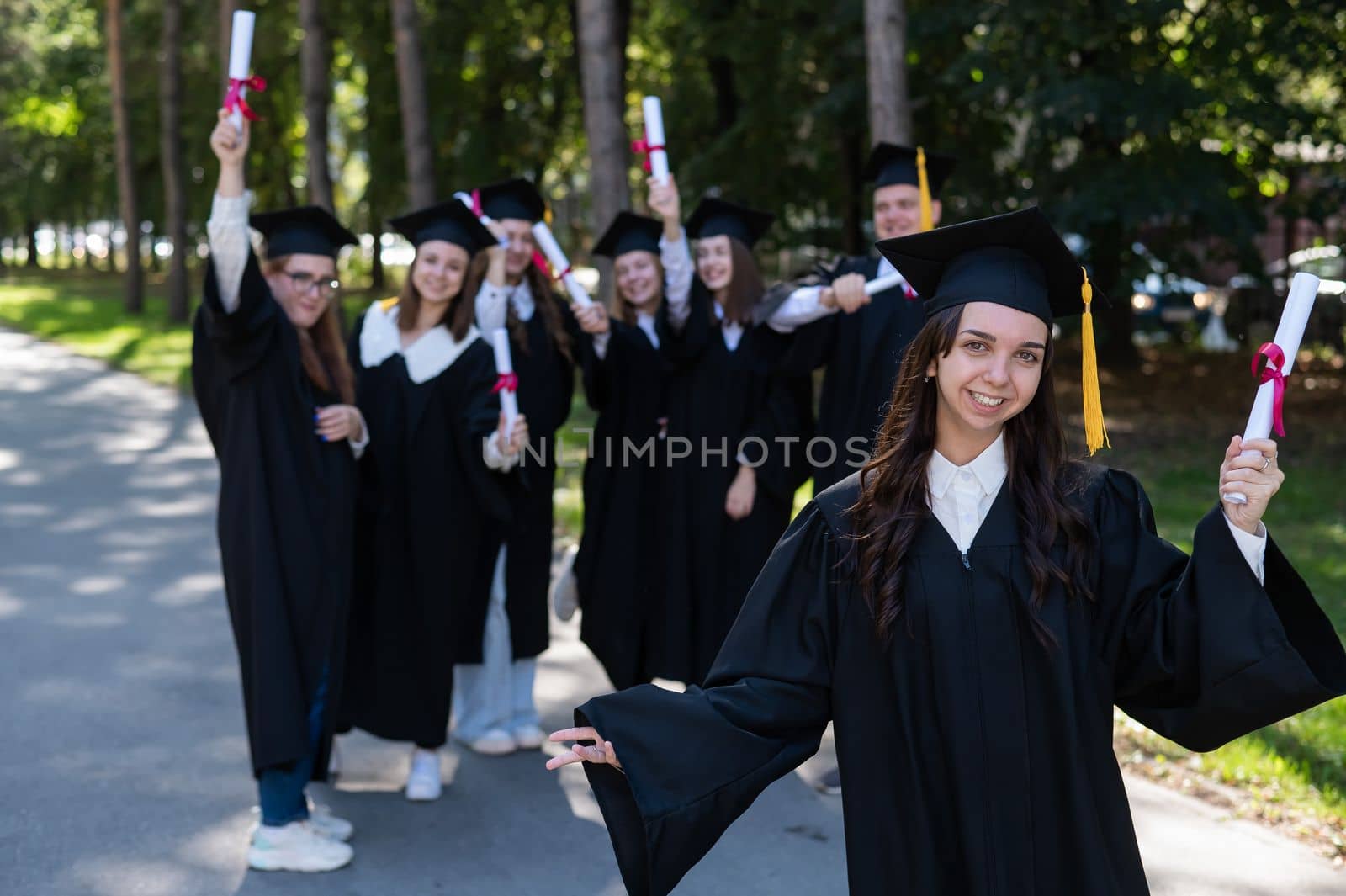 Group of happy students in graduation gowns outdoors. A young girl with a diploma in her hands in the foreground. by mrwed54