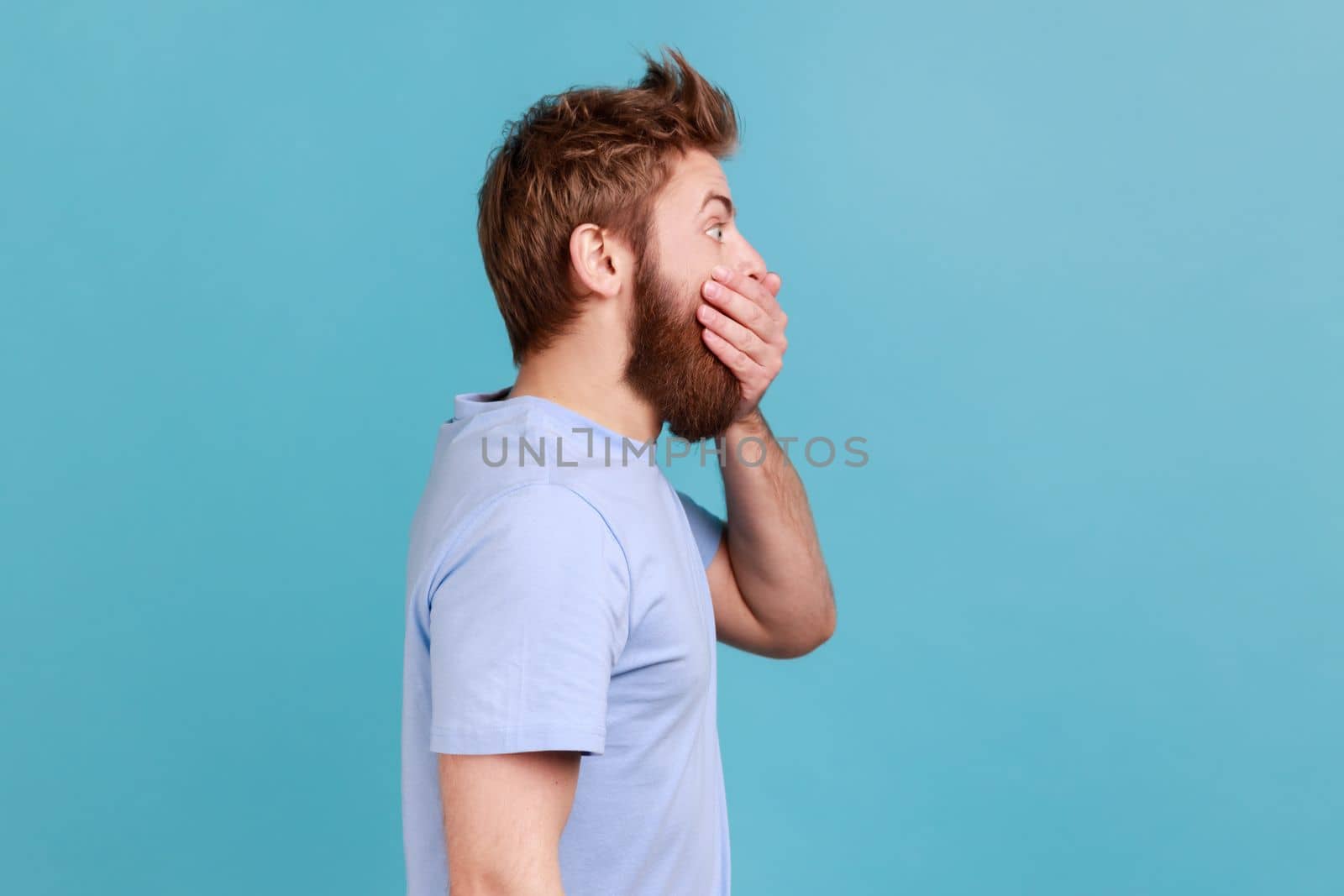 I don't tell anyone. Side view of handsome young adult bearded man zipping mouth, keeping secret information, looking at side space. Indoor studio shot isolated on blue background.