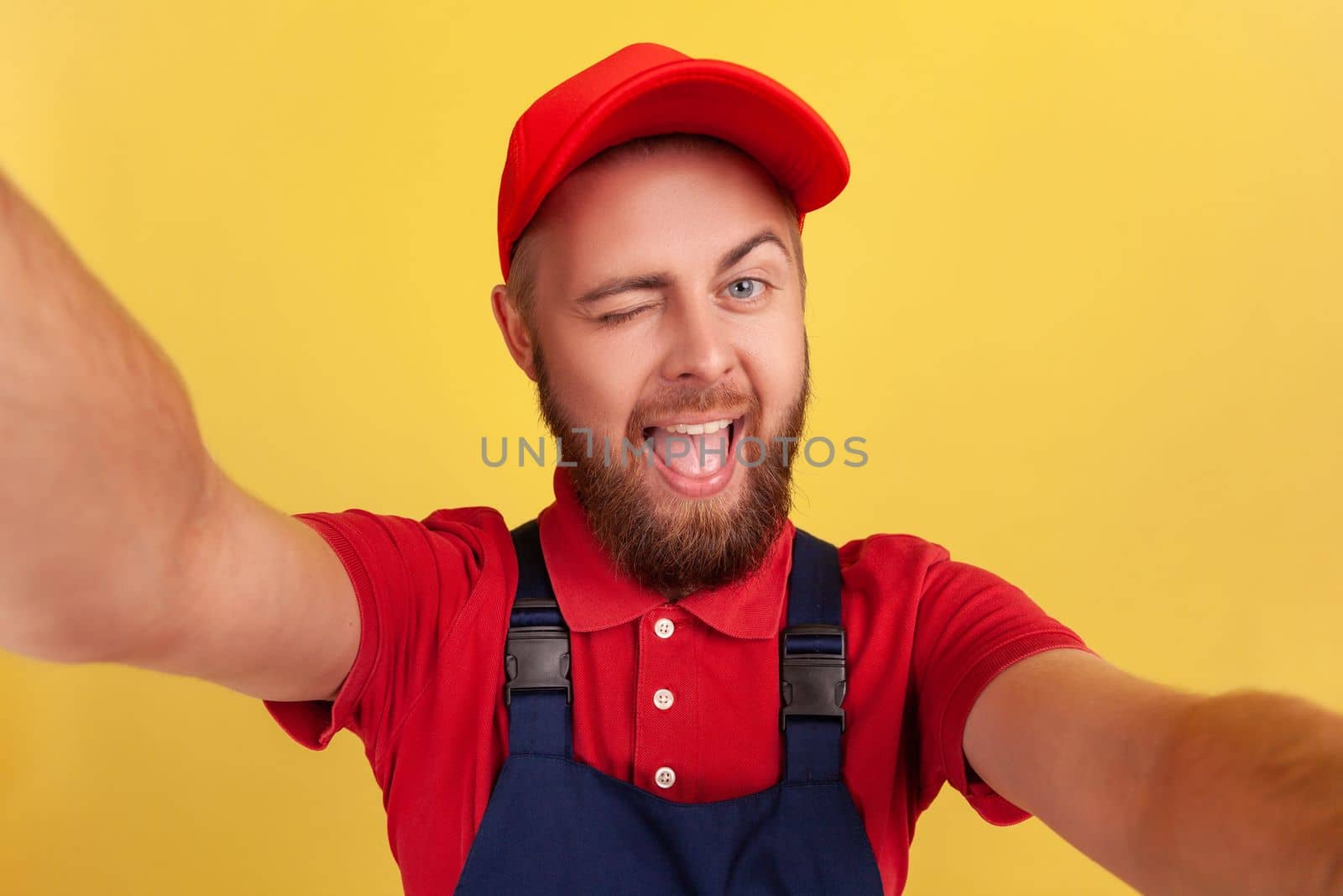 Playful delighted worker man wearing blue uniform taking selfie, looking at camera winking and showing tongue out, point of view of photo. Indoor studio shot isolated on yellow background.