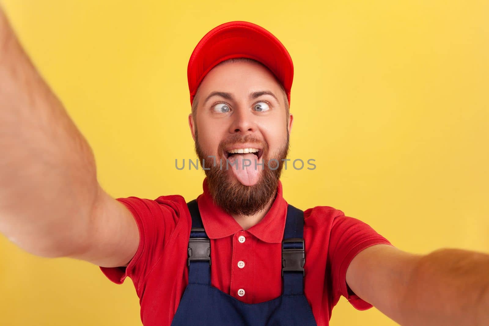 Funny handyman wearing blue uniform taking selfie, looking at camera with crossed eyes and showing tongue out POV, point of view of photo. Indoor studio shot isolated on yellow background.