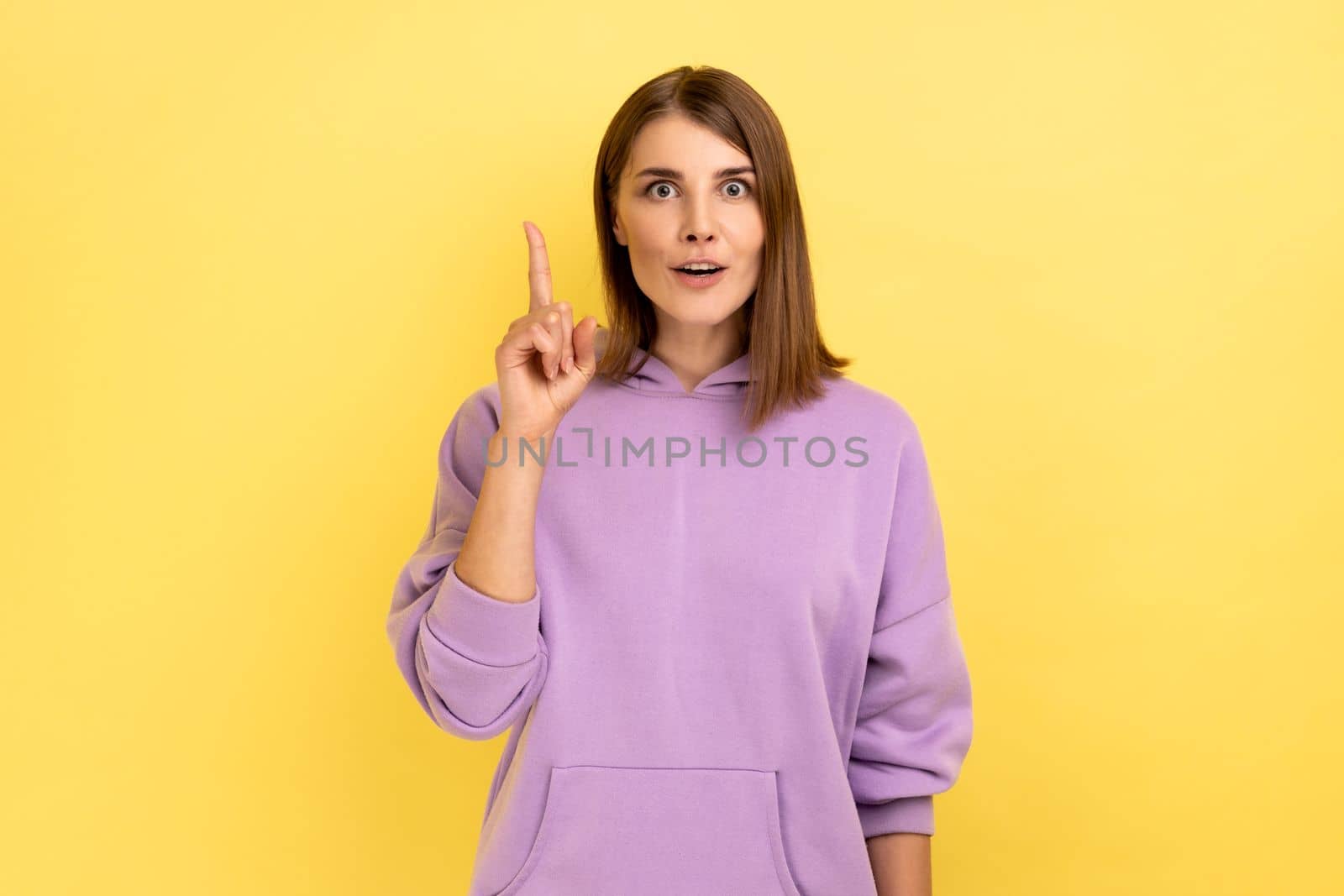 Portrait of inspired woman raising finger and having genius idea, looking amazed by sudden smart thought, wearing purple hoodie. Indoor studio shot isolated on yellow background.