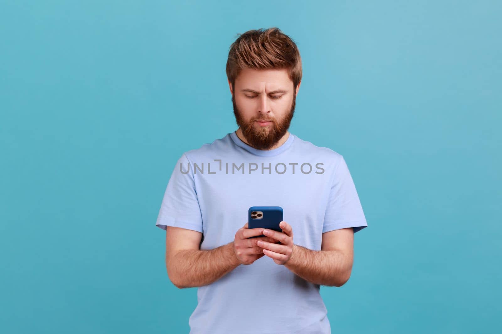 Portrait of serious concentrated bearded man texting message in social media on cell phone, using mobile network services, chatting online. Indoor studio shot isolated on blue background.