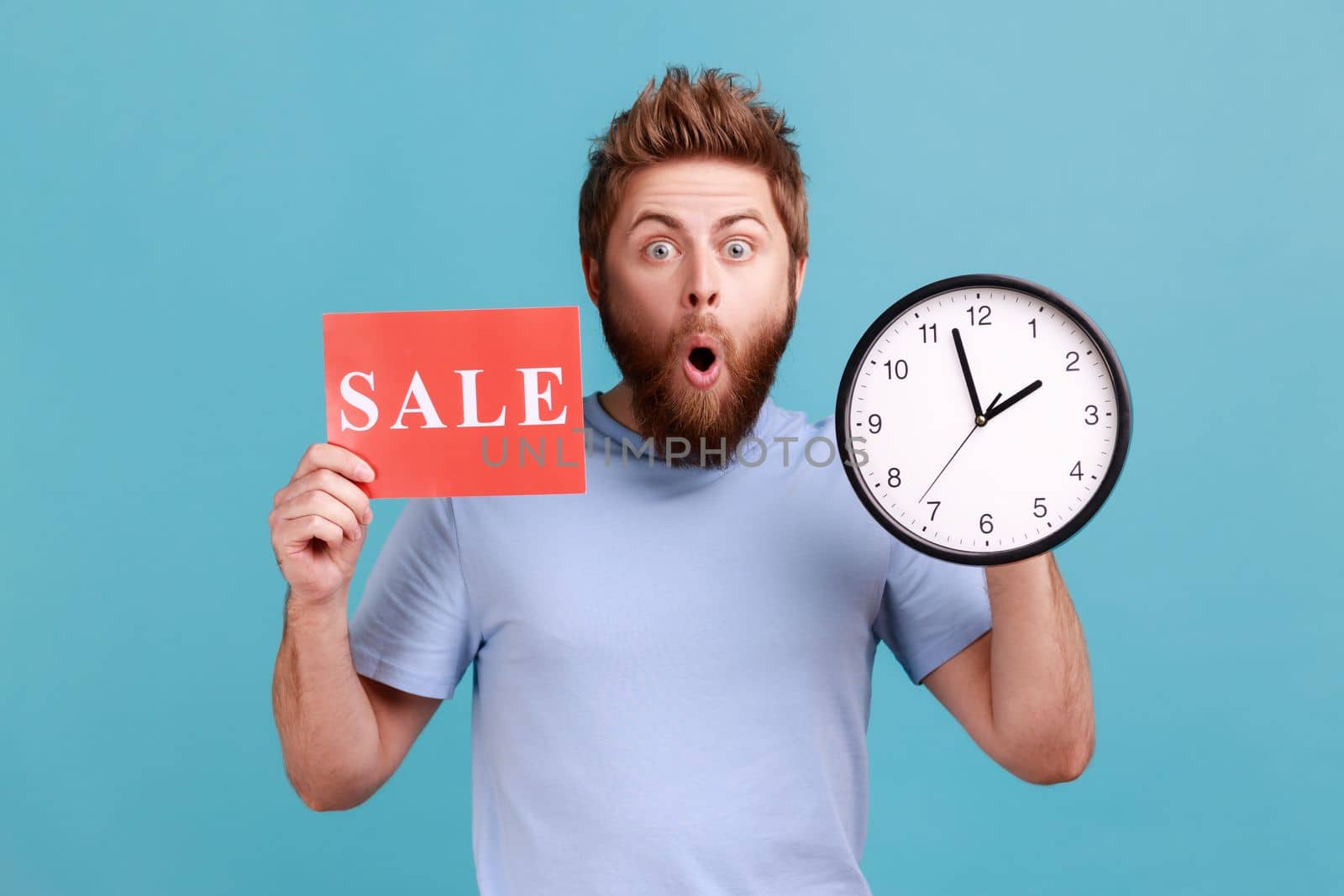 Portrait of shocked astonished handsome bearded man holding clock and card with sale inscription, announcing finishing huge discounts. Indoor studio shot isolated on blue background.