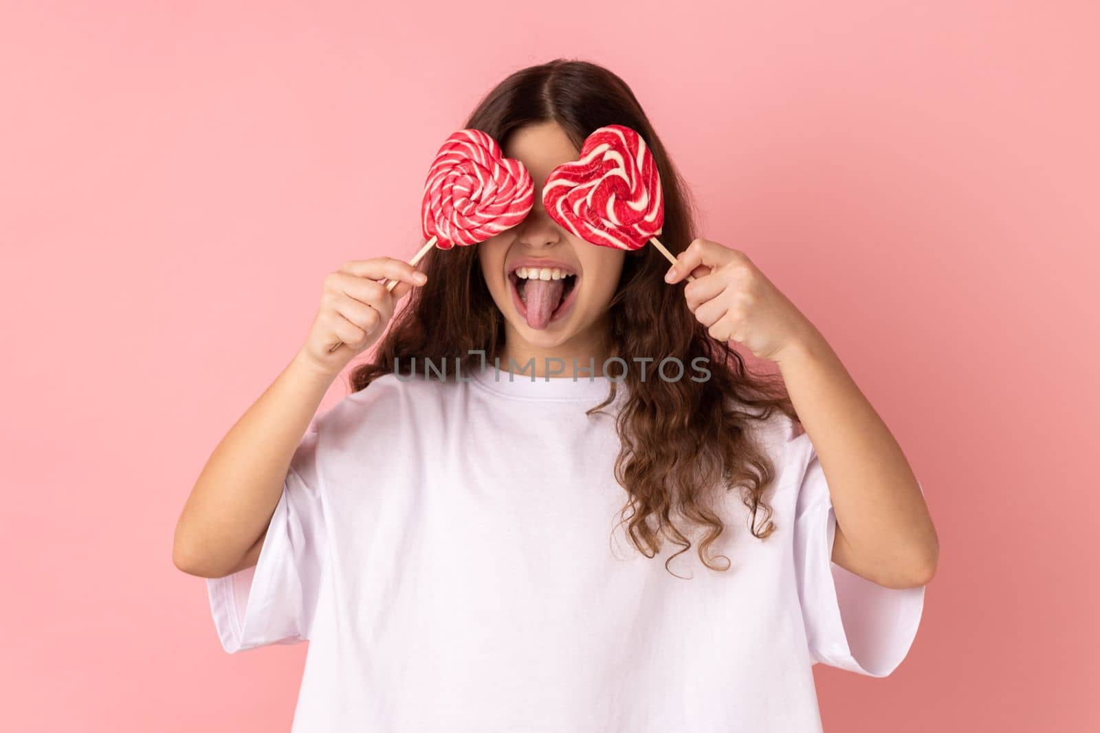 Portrait of little girl wearing white T-shirt covering her eyes with heart shape candies and showing tongue out, posing with happy expression. Indoor studio shot isolated on pink background.