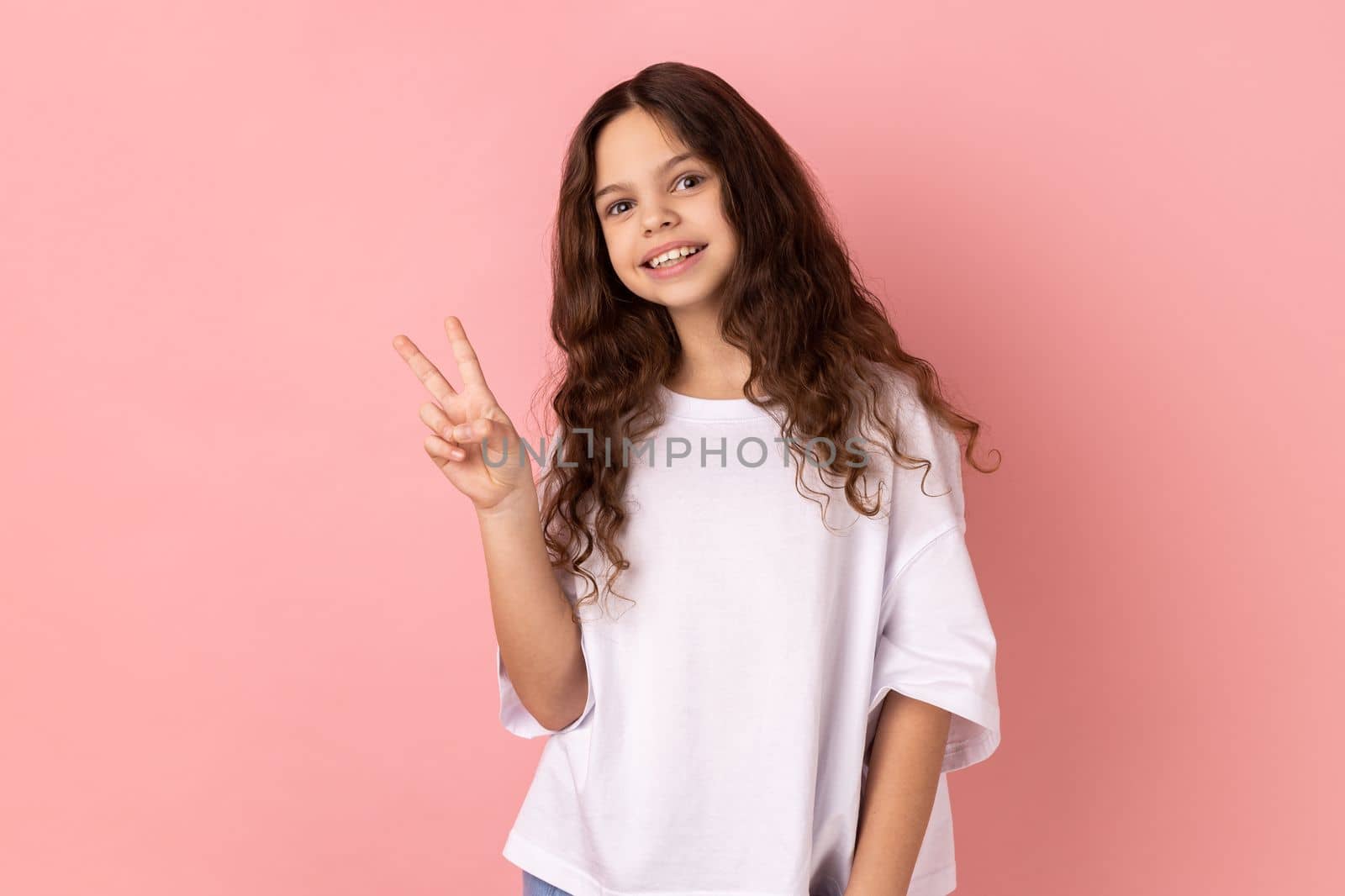 Portrait of positive optimistic little girl wearing white T-shirt looking at camera with toothy smile, showing v sign, peace gesture. Indoor studio shot isolated on pink background.
