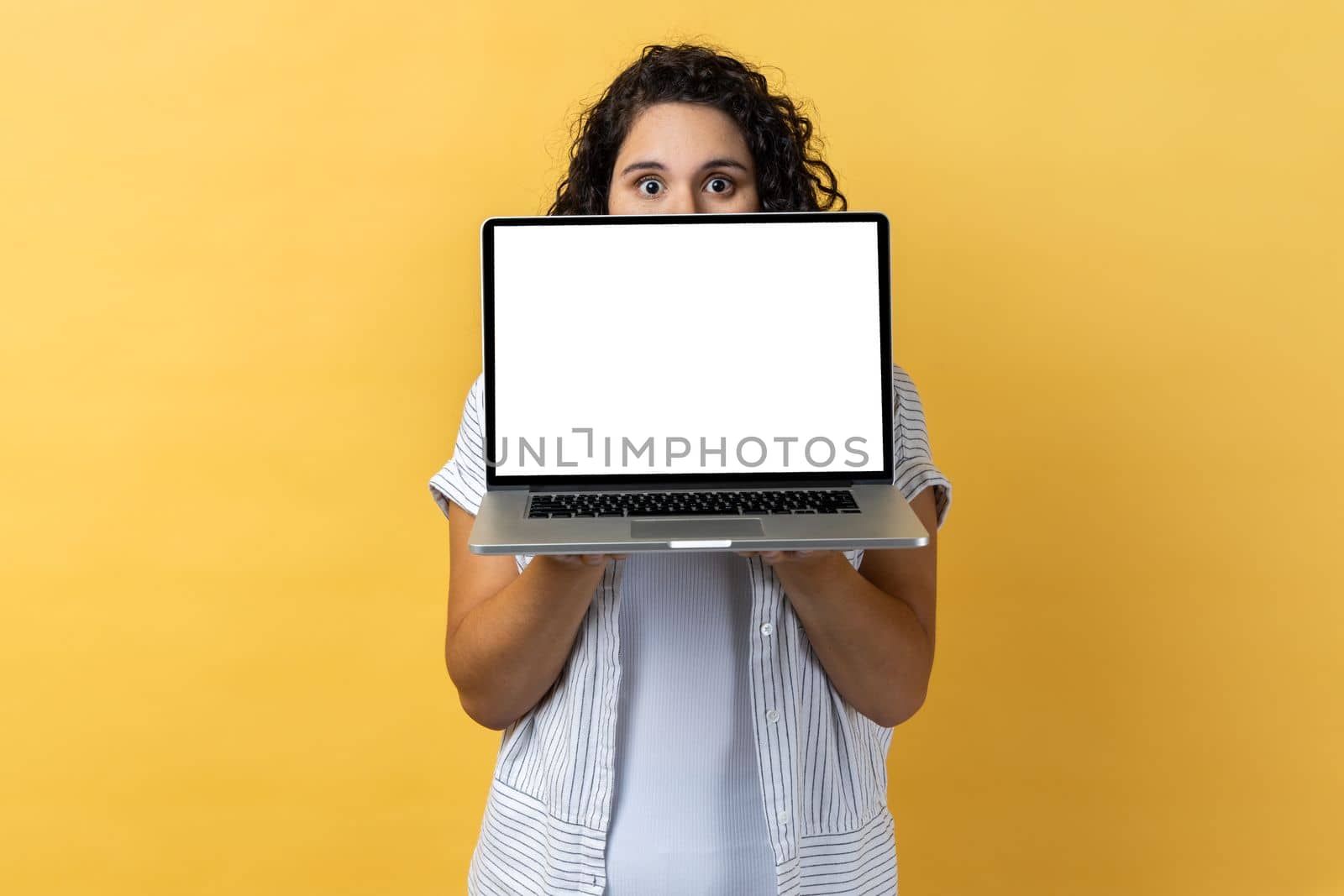 Portrait of shy woman with dark wavy hair hiding half of her face behind laptop with empty display, mockup, copy space for promotion. Indoor studio shot isolated on yellow background.