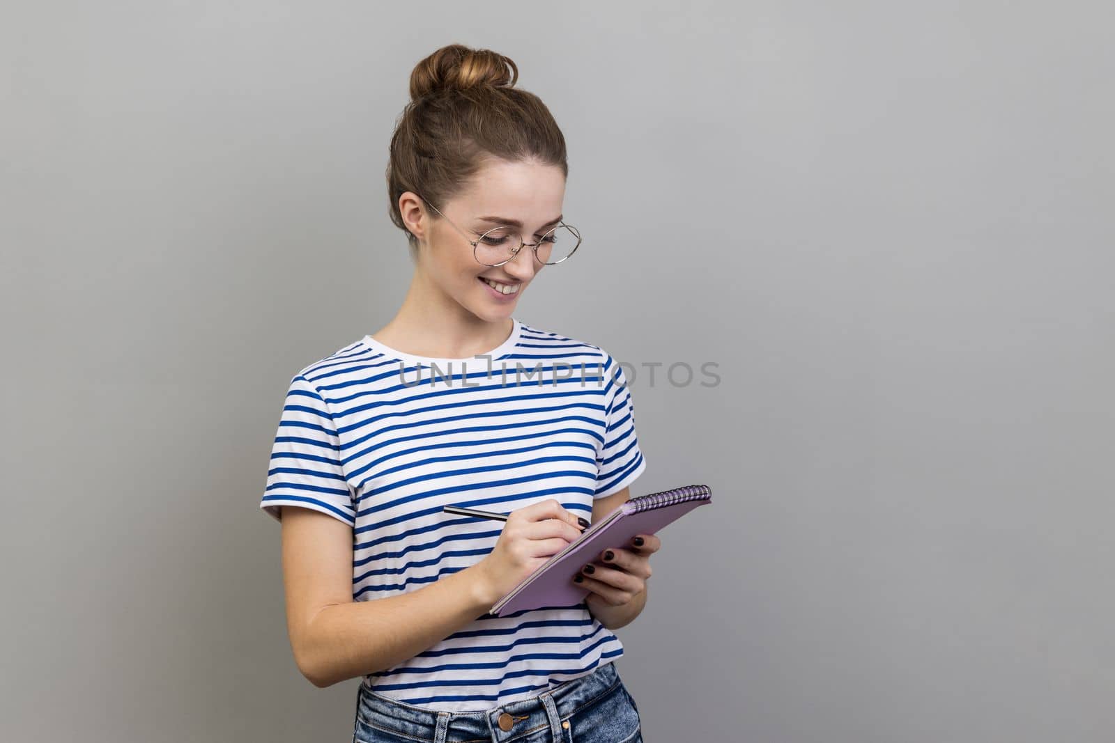 Portrait of happy inspired woman wearing striped T-shirt and glasses writing down interesting idea in notebook, making plans, to-do list. Indoor studio shot isolated on gray background.