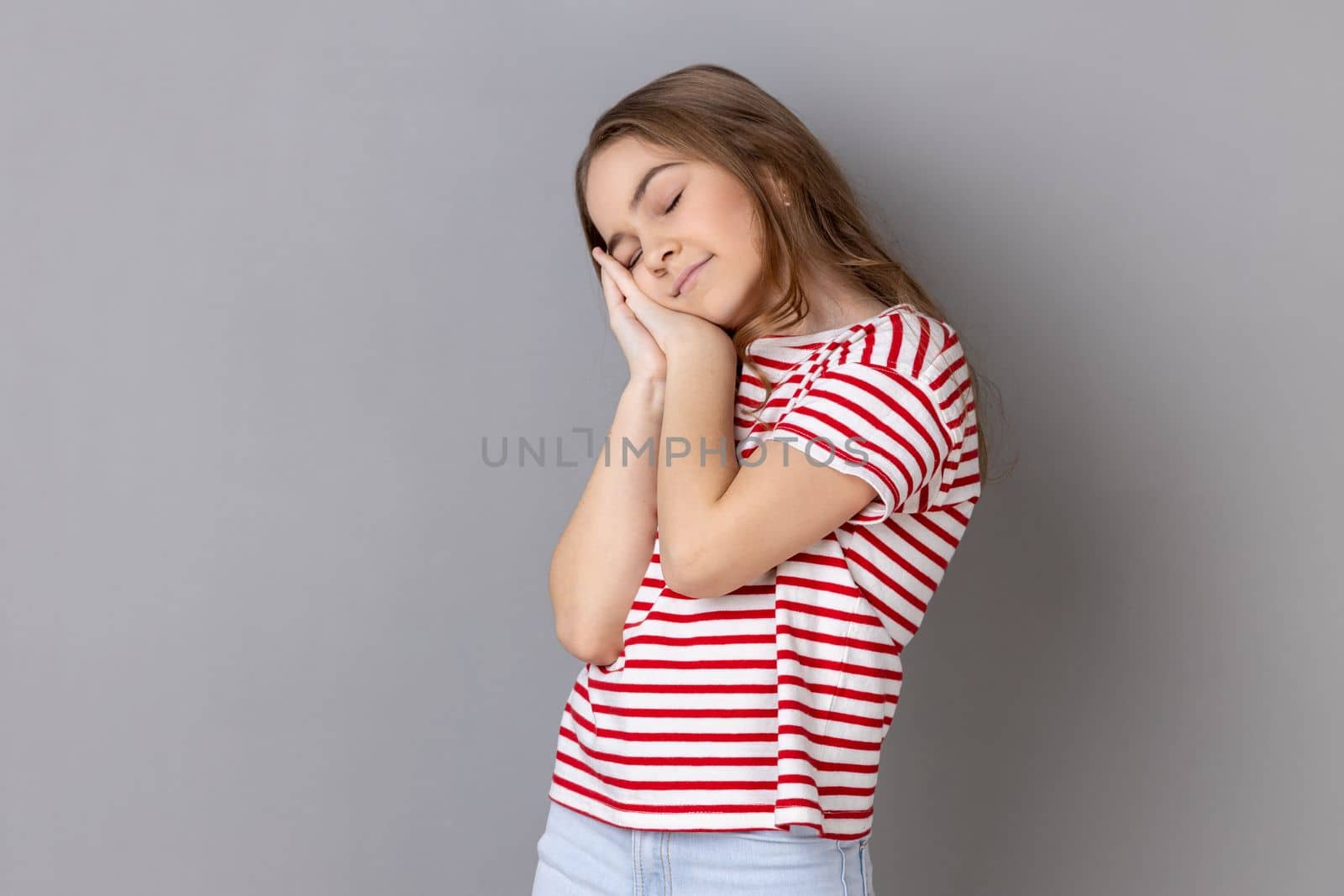 I need rest. Portrait of little girl wearing striped T-shirt yawning and covering mouth with hand, feeling exhausted, lack of sleep. Indoor studio shot isolated on gray background.