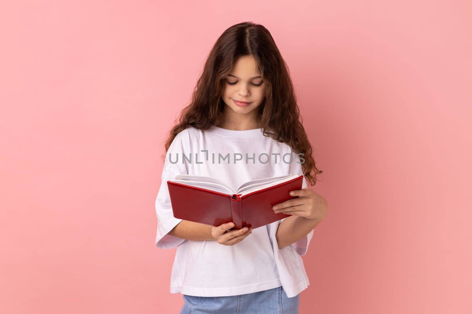 Portrait of delighted charming little girl wearing white T-shirt holding in hands and reading interesting book, being very attentive. Indoor studio shot isolated on pink background.