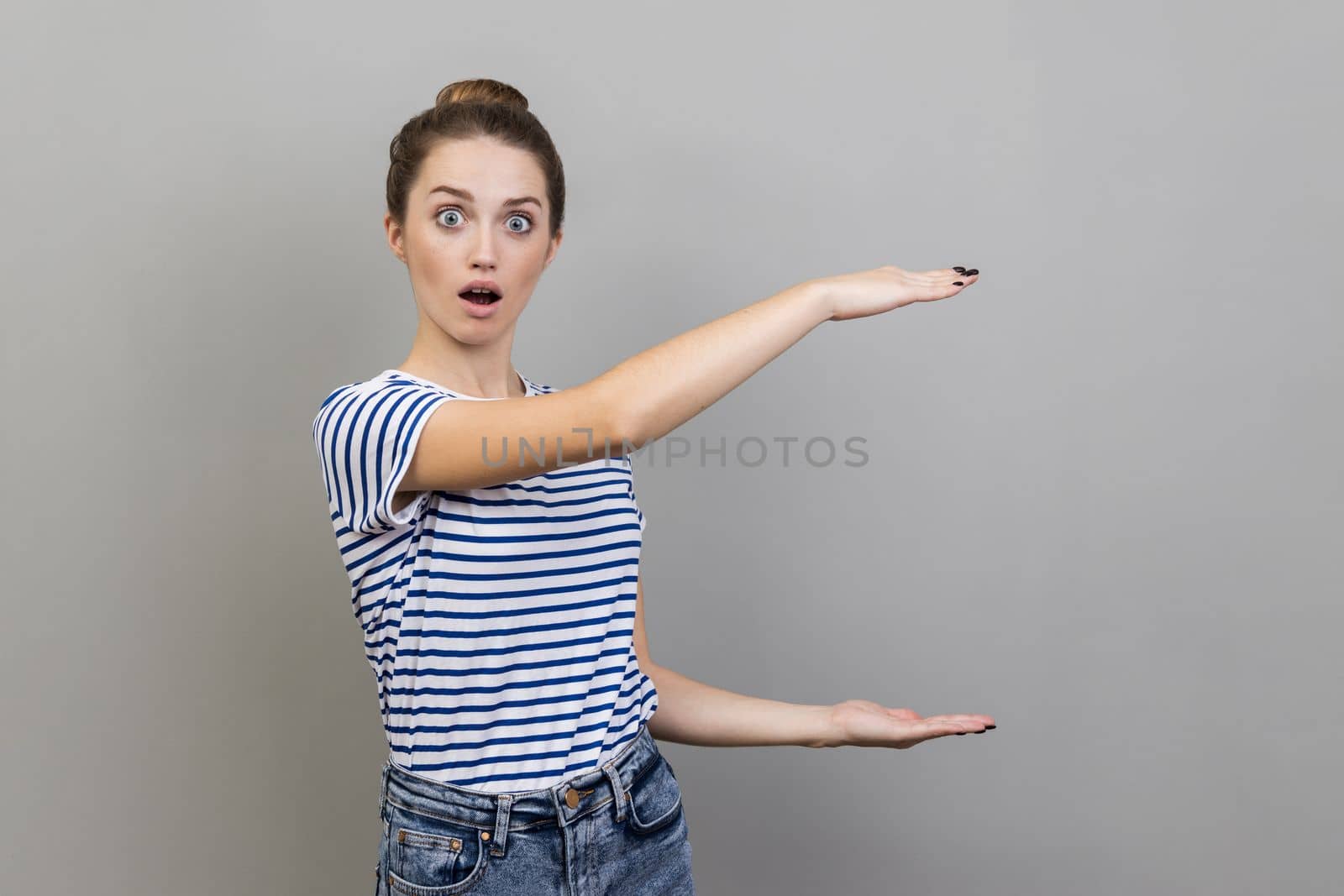 Portrait of woman wearing striped T-shirt showing big empty space, holding box copy space, keeps mouth open, looking at camera with shocked expression. Indoor studio shot isolated on gray background.
