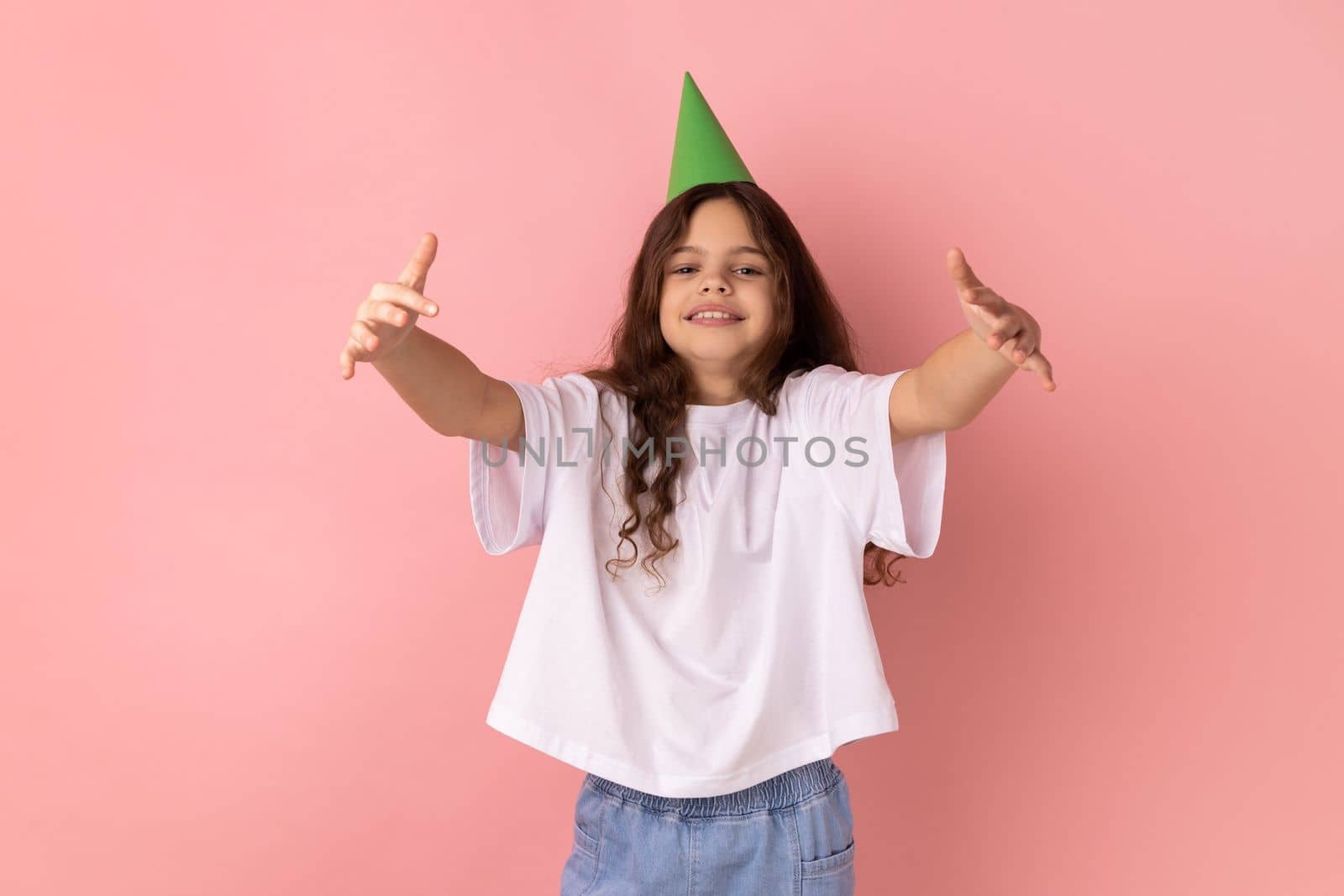 Portrait of delighted little girl wearing white T-shirt and party cone giving free hugs with outstretched hands, welcoming inviting to embrace. Indoor studio shot isolated on pink background.