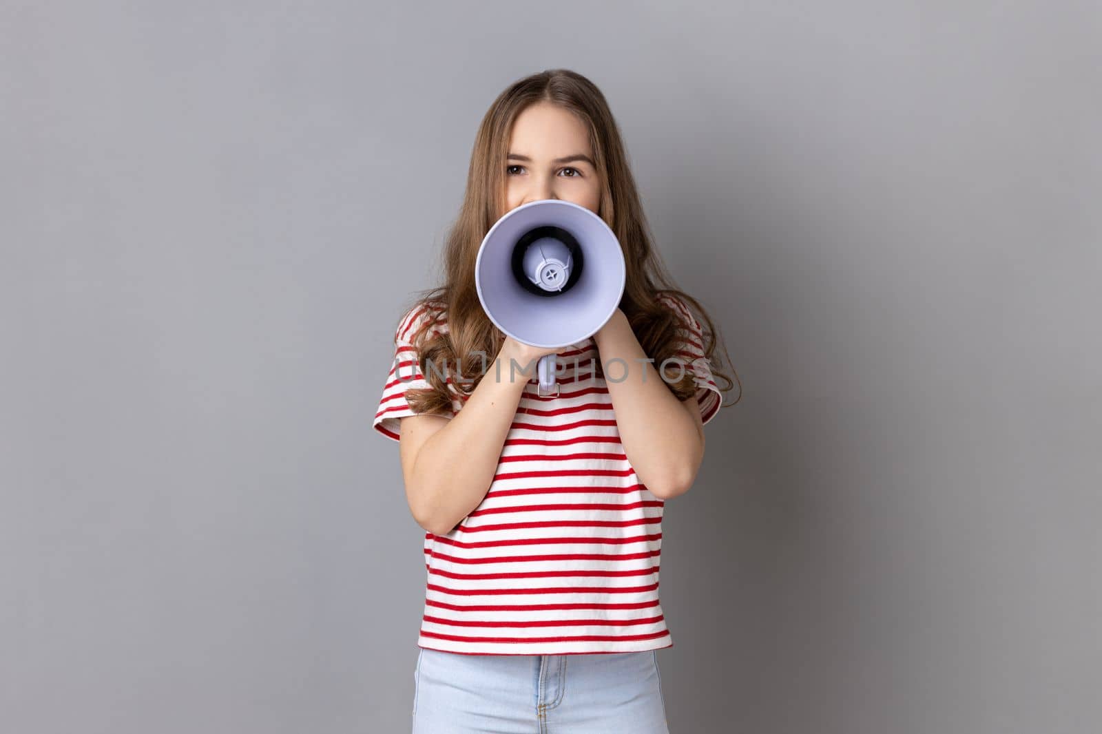 Portrait of dark haired little girl wearing striped T-shirt screaming in megaphone, speaking, announcing important information. Indoor studio shot isolated on gray background.