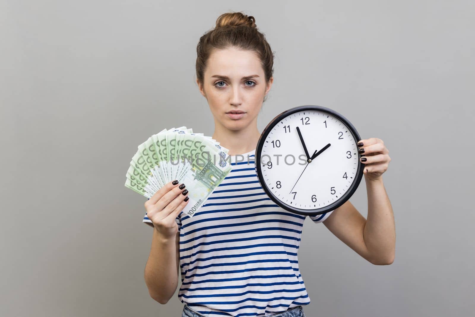 Portrait of strict serious woman with bun hairstyle wearing striped T-shirt holding wall clock and fun of euro banknotes, looking at camera. Indoor studio shot isolated on gray background.