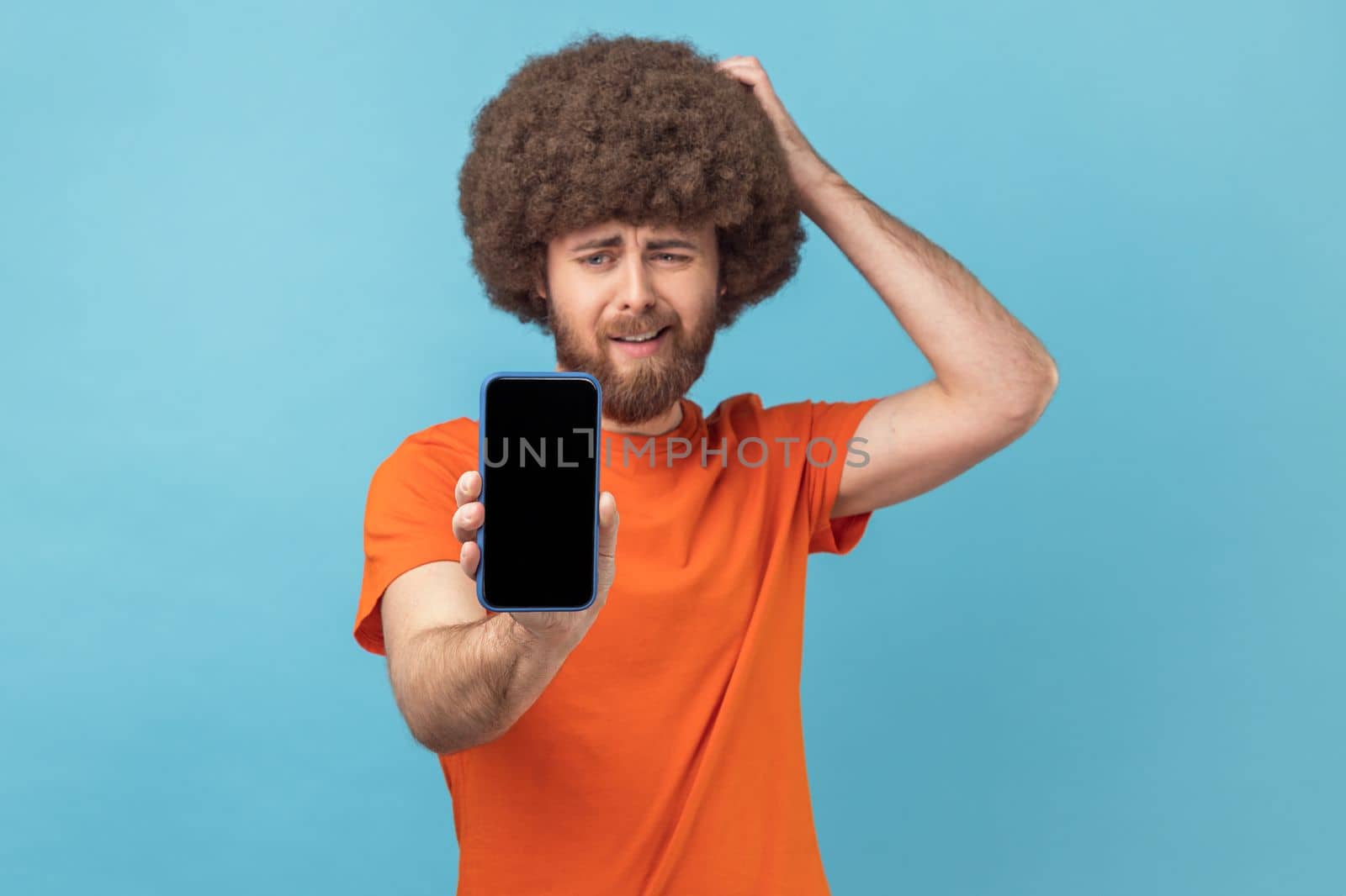 Portrait of puzzled pensive man with Afro hairstyle wearing orange T-shirt showing cell phone with empty display, presenting area for advertisement. Indoor studio shot isolated on blue background.