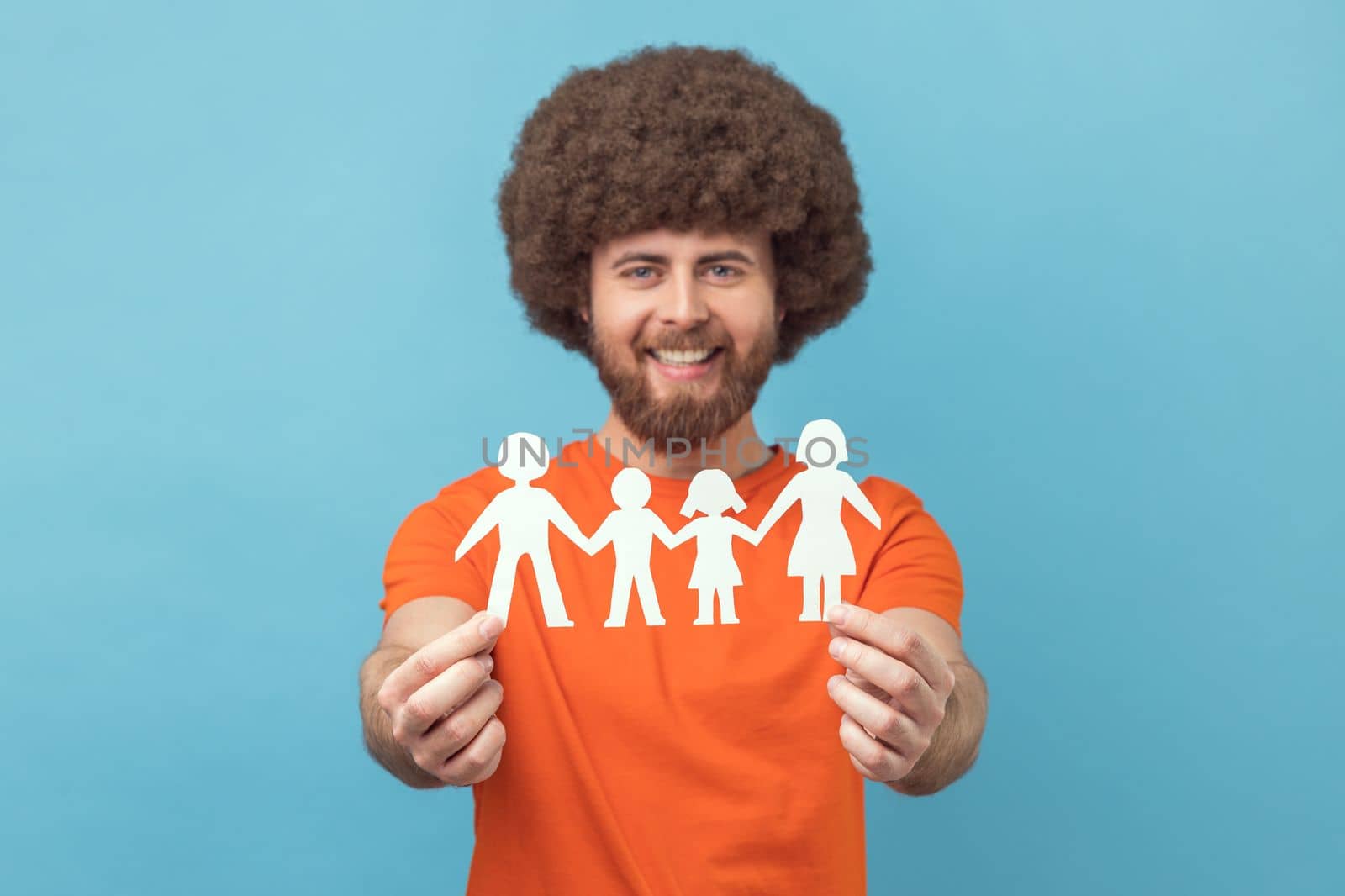 Portrait of smiling satisfied man with Afro hairstyle wearing orange T-shirt holding paper symbols of family, looking at camera, relationship. Indoor studio shot isolated on blue background.