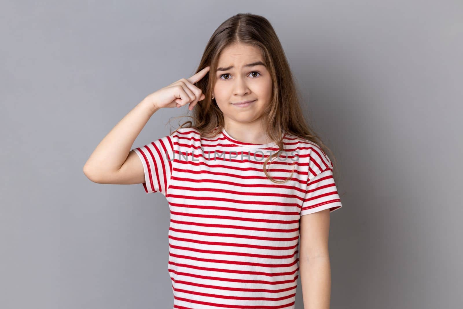 Are you kidding me. Portrait of little girl wearing striped T-shirt holding finger near head temple and gesturing, looking displeased with crazy idea. Indoor studio shot isolated on gray background.