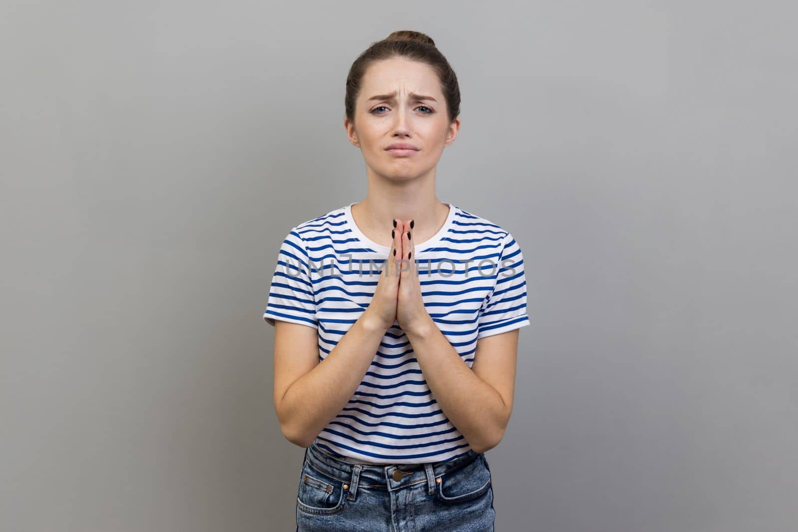 Please, I need. Portrait of woman wearing striped T-shirt holding hands in prayer and asking for help with pleading imploring expression, full of hope. Indoor studio shot isolated on gray background.