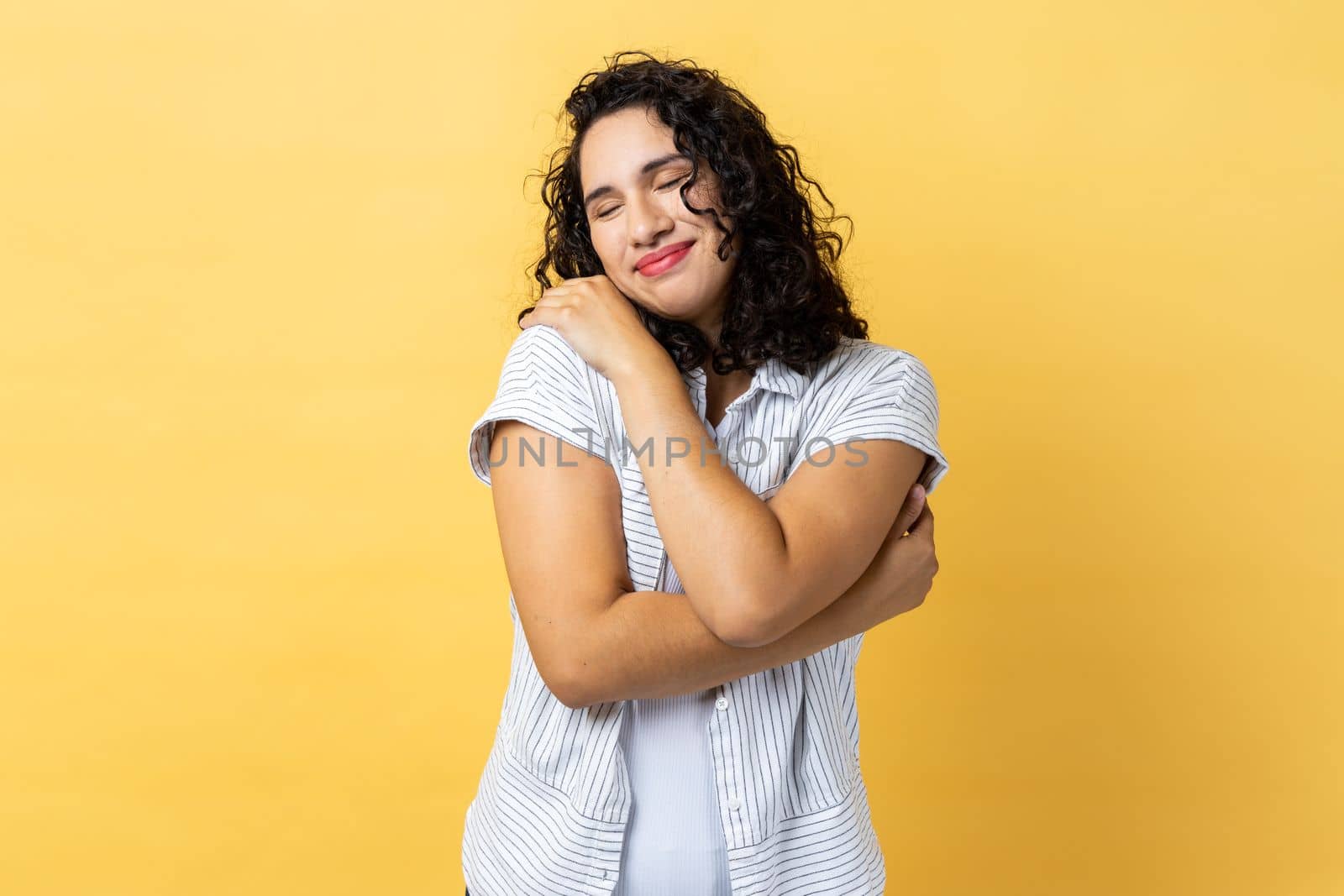 Portrait of delighted woman with dark wavy hair hugging herself and smiling with dreamy satisfied expression, love myself gesture. Indoor studio shot isolated on yellow background.