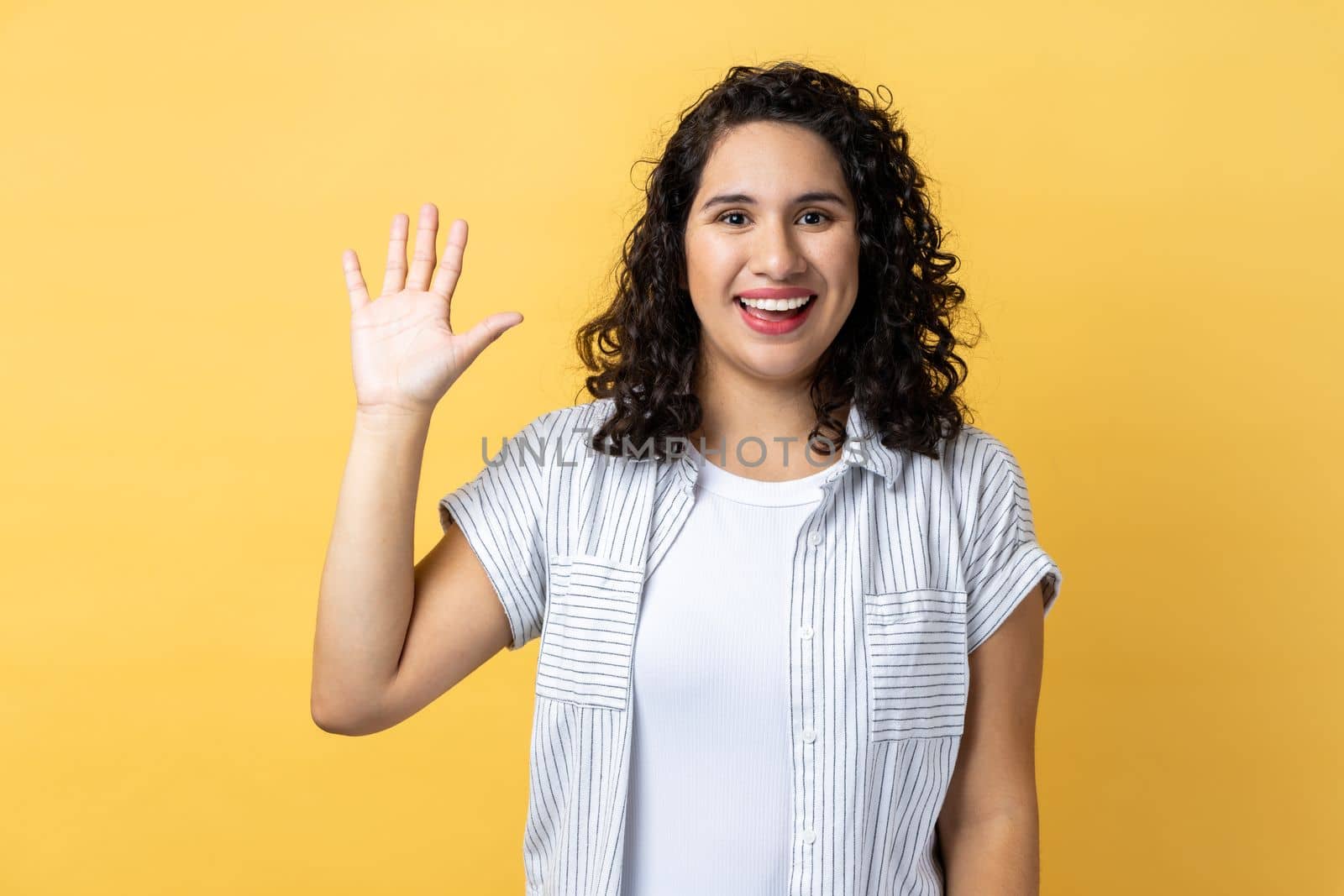 Portrait of friendly positive beautiful woman with dark wavy hair smiles toothily, raises palm greets friend, saying hello or goodbye. Indoor studio shot isolated on yellow background.