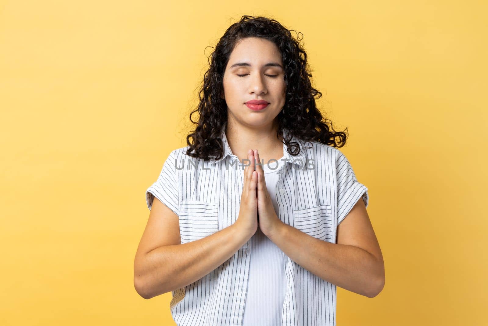 Portrait of peaceful calm relaxed woman with dark wavy hair standing with prayer gesture, keeping eyes closed, meditating sitting in yoga position. Indoor studio shot isolated on yellow background.