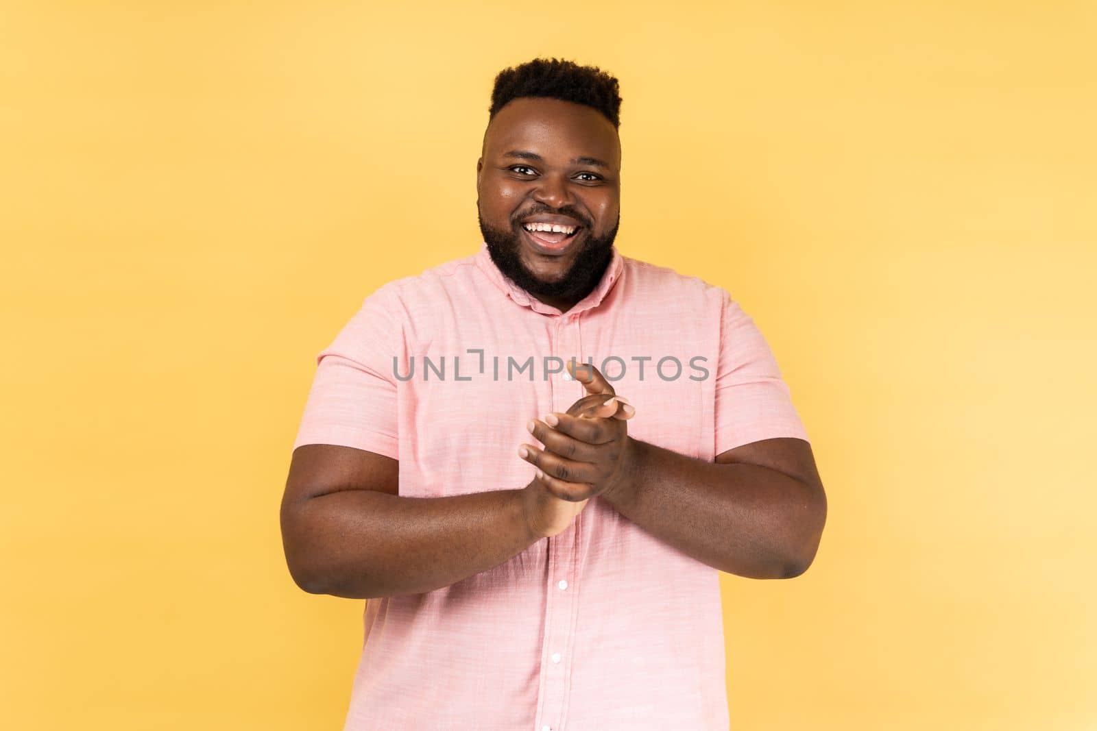 Portrait of positive friendly man wearing pink shirt standing with palms together, applauding, clapping hands congratulating you with victory. Indoor studio shot isolated on yellow background.