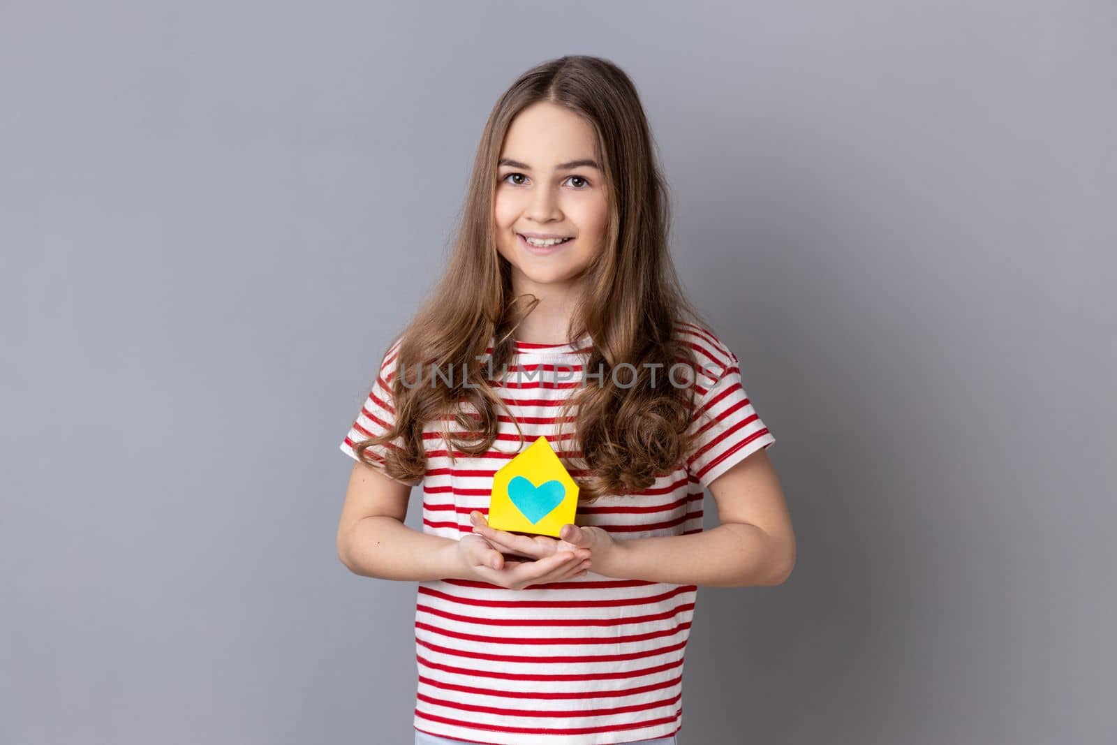Portrait of little girl wearing striped T-shirt holding in hands little paper house looking at camera with smile, dreaming about apartment. Indoor studio shot isolated on gray background.