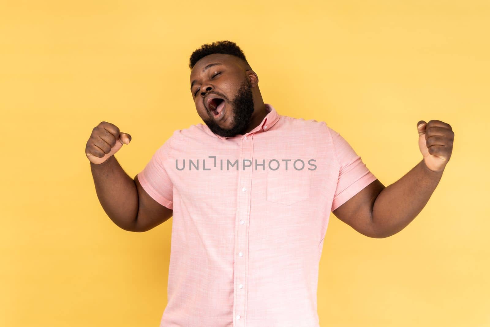 Portrait of tired bearded man wearing pink shirt yawning, feeling exhausted, lack of sleep, outstretching arms, keeps eyes closed. Indoor studio shot isolated on yellow background.