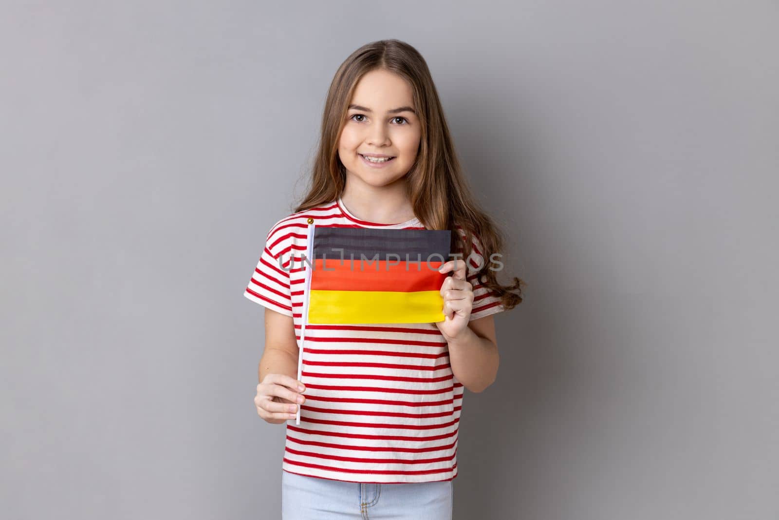 Germany flag. Portrait of satisfied optimistic adorable cute little girl wearing striped T-shirt holding german flag, looking at camera. Indoor studio shot isolated on gray background.