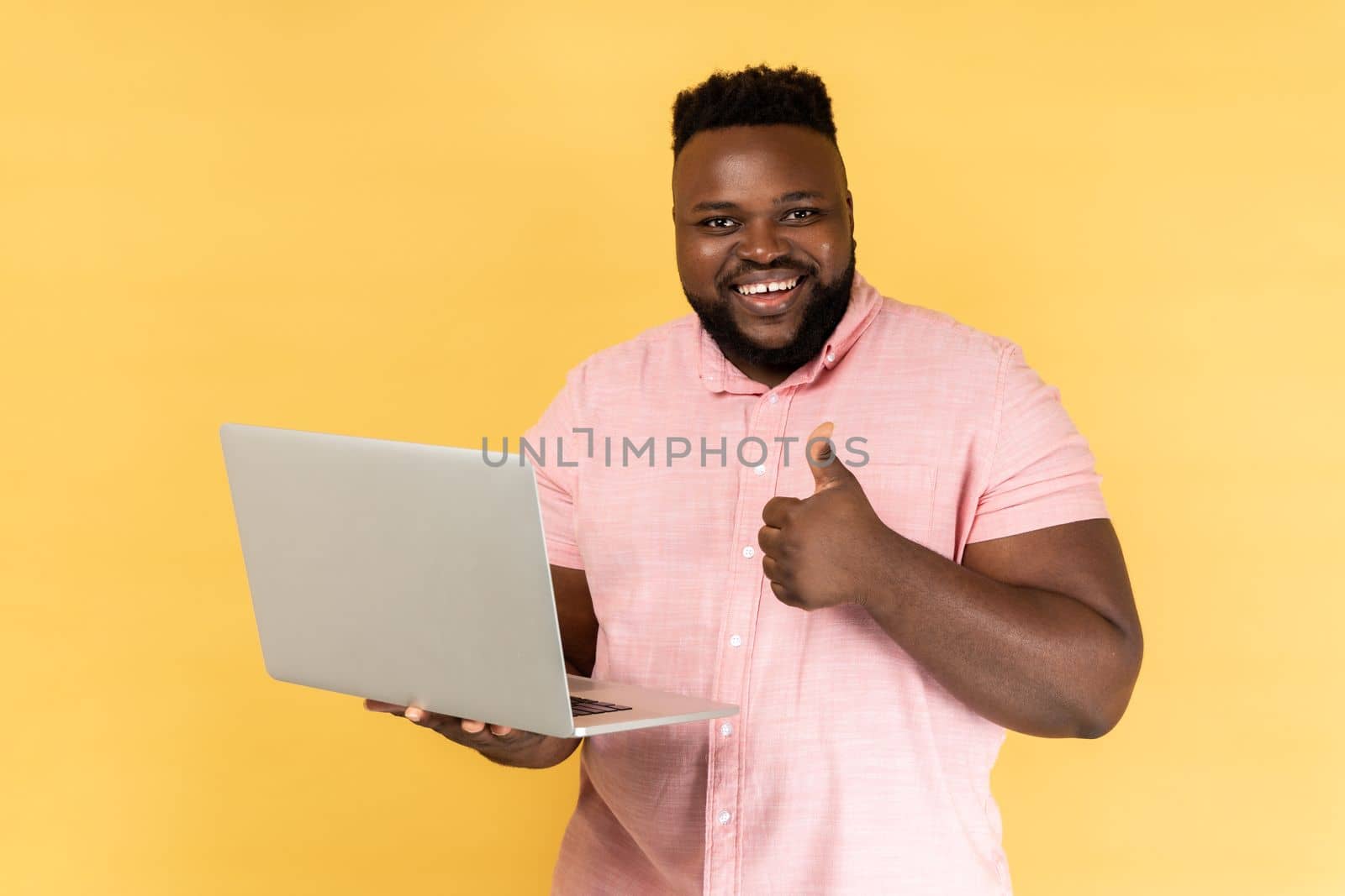 Portrait of smiling man holding laptop in hand showing thumbs up gesture, blogger likes posts, looking at camera, recommending to followers. Indoor studio shot isolated on yellow background.