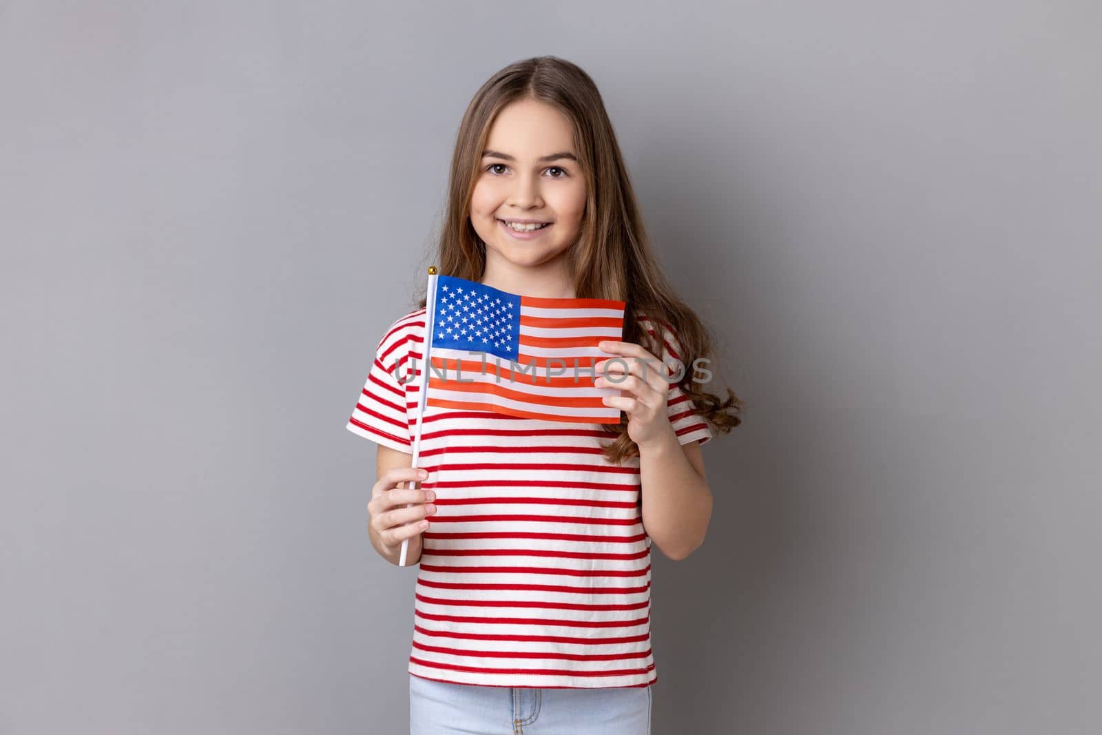 American flag. Portrait of delighted happy adorable cute little girl wearing striped T-shirt holding flag of United States of America, looking at camera. Indoor studio shot isolated on gray background