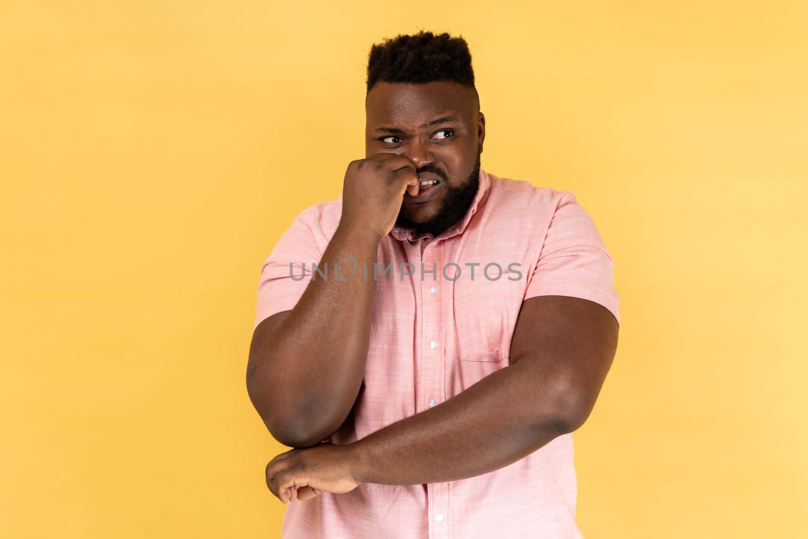 Anxiety disorder. Portrait of stressed out worried man in pink shirt biting nails, nervous about troubles, panicking and looking scared, looking away. Indoor studio shot isolated on yellow background.