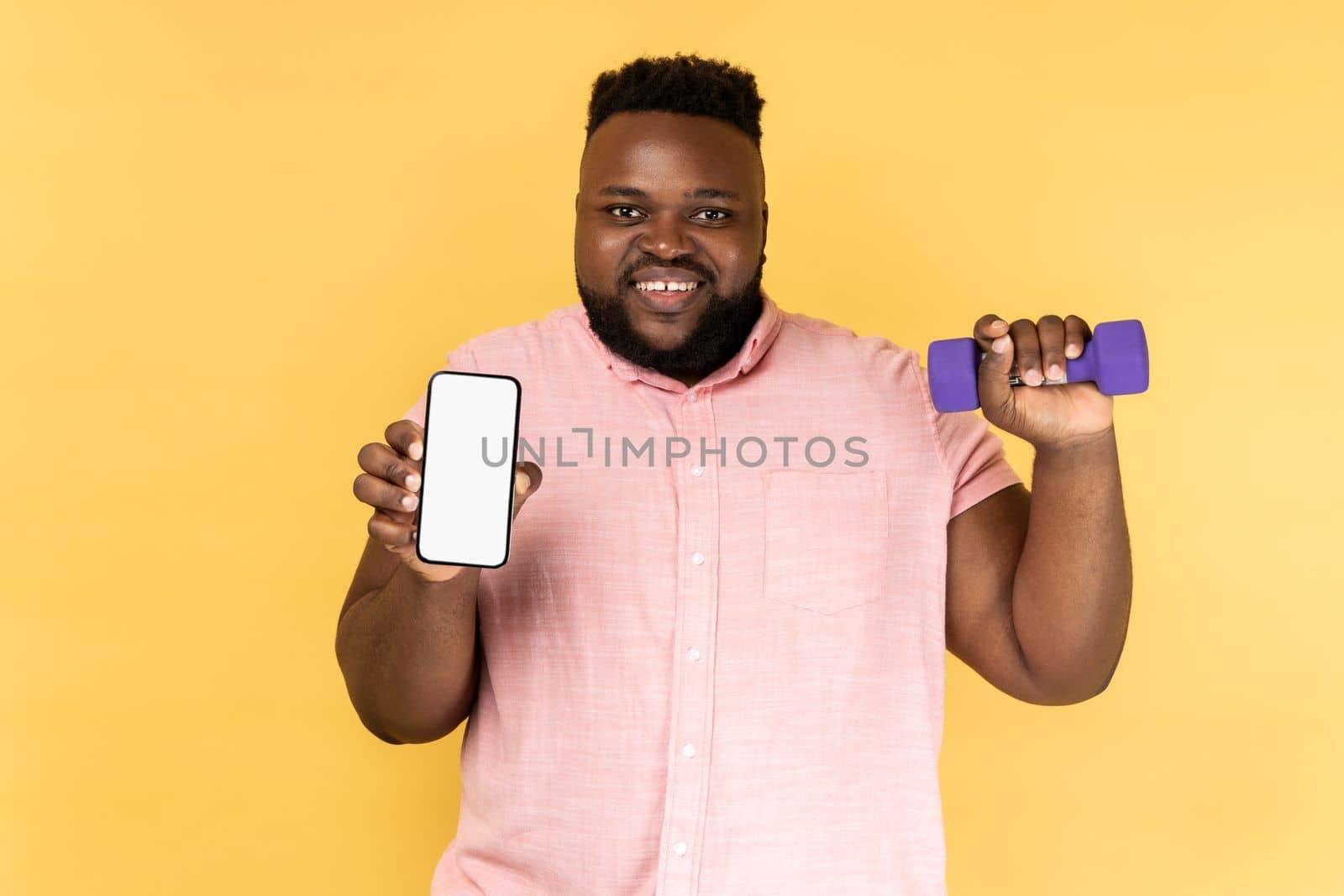 Portrait of happy joyful bearded man wearing pink shirt standing holding dumbbell and smart phone with empty display for advertisement. Indoor studio shot isolated on yellow background.