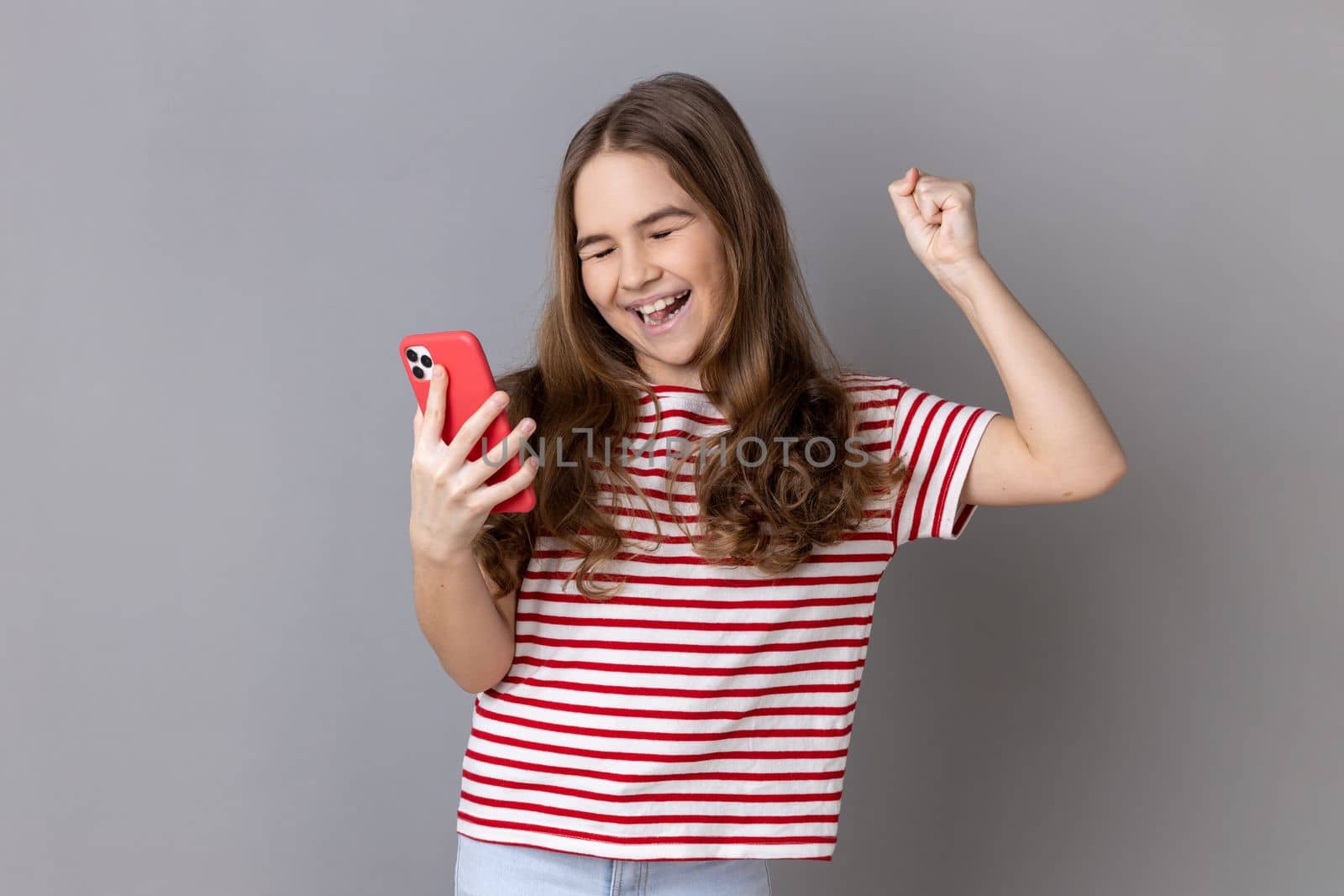 Portrait of excited amazed little girl wearing striped T-shirt standing holding phone, looking at camera and celebrate victory with fist. Indoor studio shot isolated on gray background.
