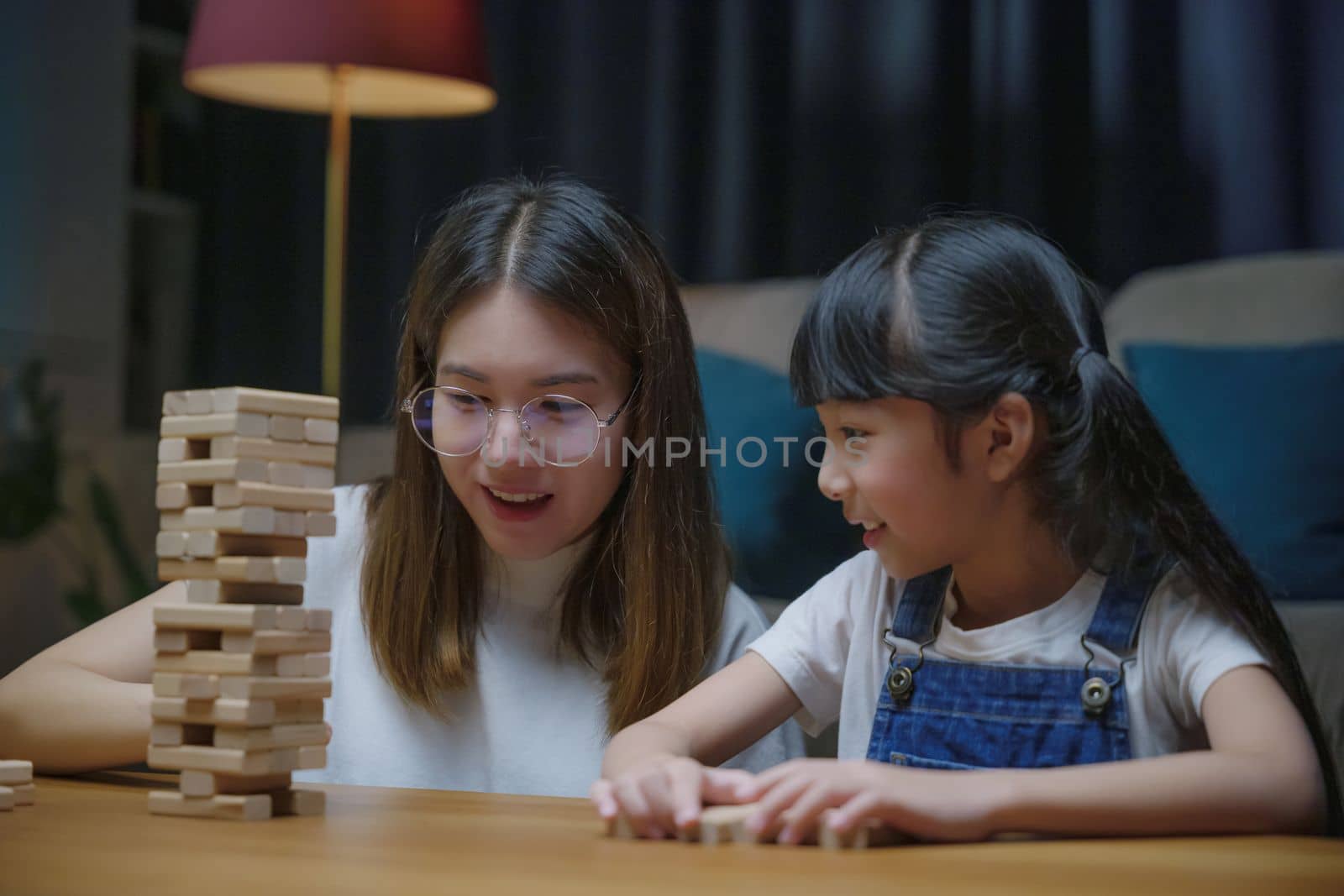 Asian young mother playing game in wood block with little daughter in home living room at night, Smiling woman help teach preschooler kid play build constructor tower of wooden blocks, family funny