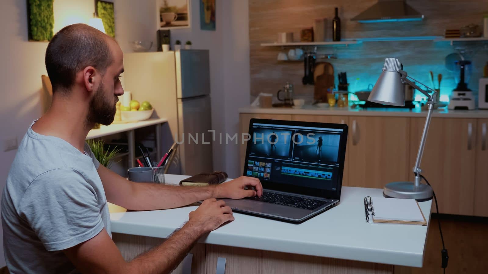 Content creator working remotely on laptop from home during night time. Videographer editing audio film montage on professional laptop sitting on desk in modern kitchen in midnight